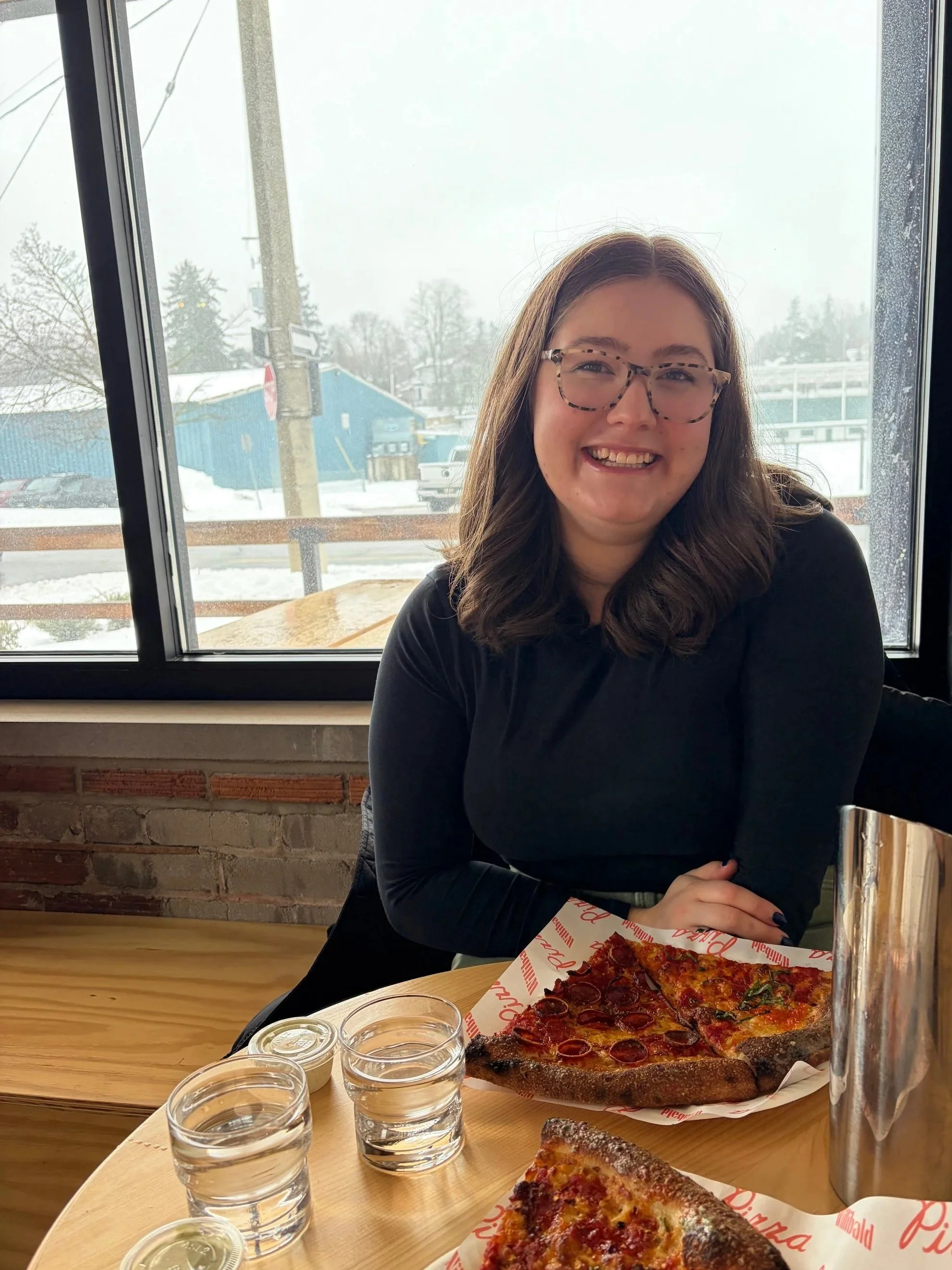 A woman with glasses smiling at a restaurant table with a pizza and several glasses of water, sitting next to a window with a snowy outdoor scene.