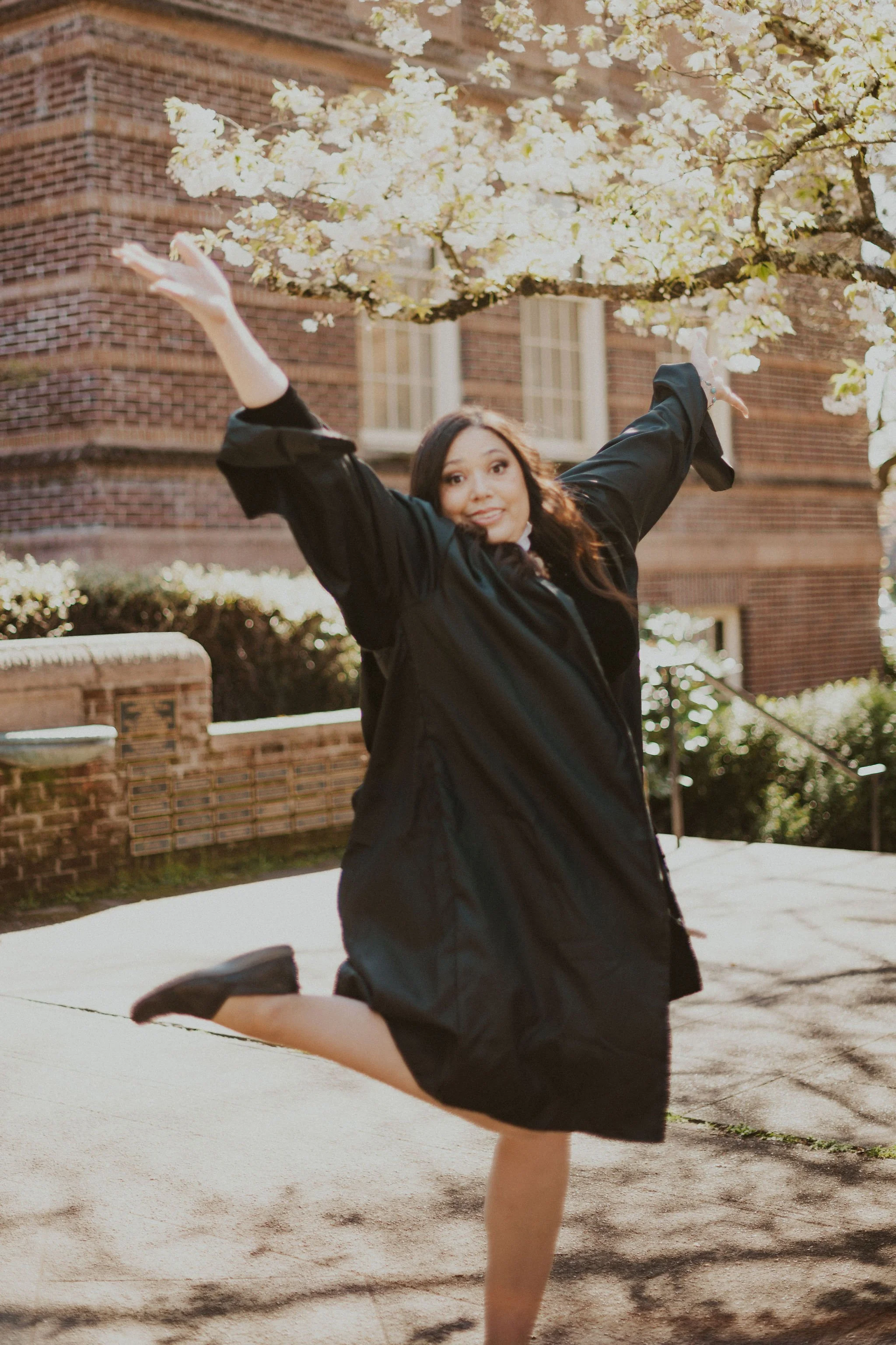 A woman in a black graduation gown is happily jumping and spinning outdoors near a blooming tree with white flowers, in front of a brick building.