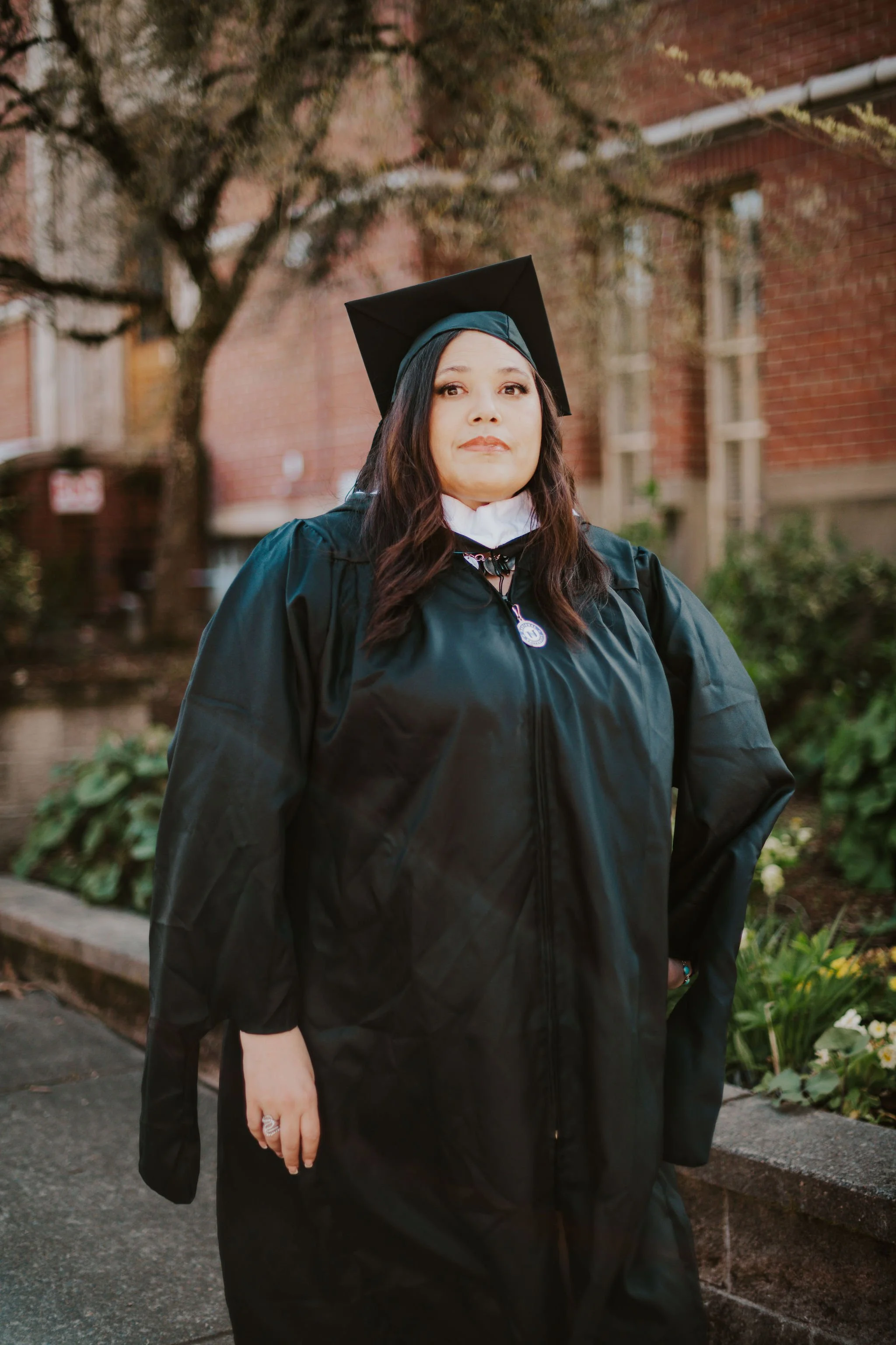 A woman in graduation cap and gown standing outdoors near a brick building and trees.