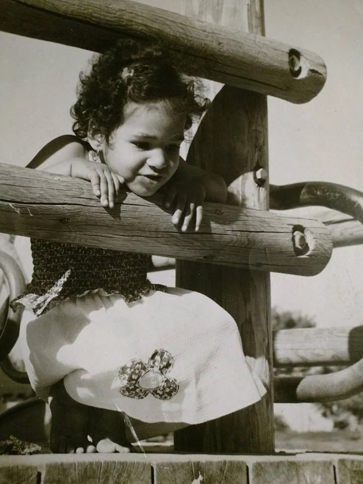 A young girl crouched behind wooden rails on a playground, smiling and peering through the gaps.