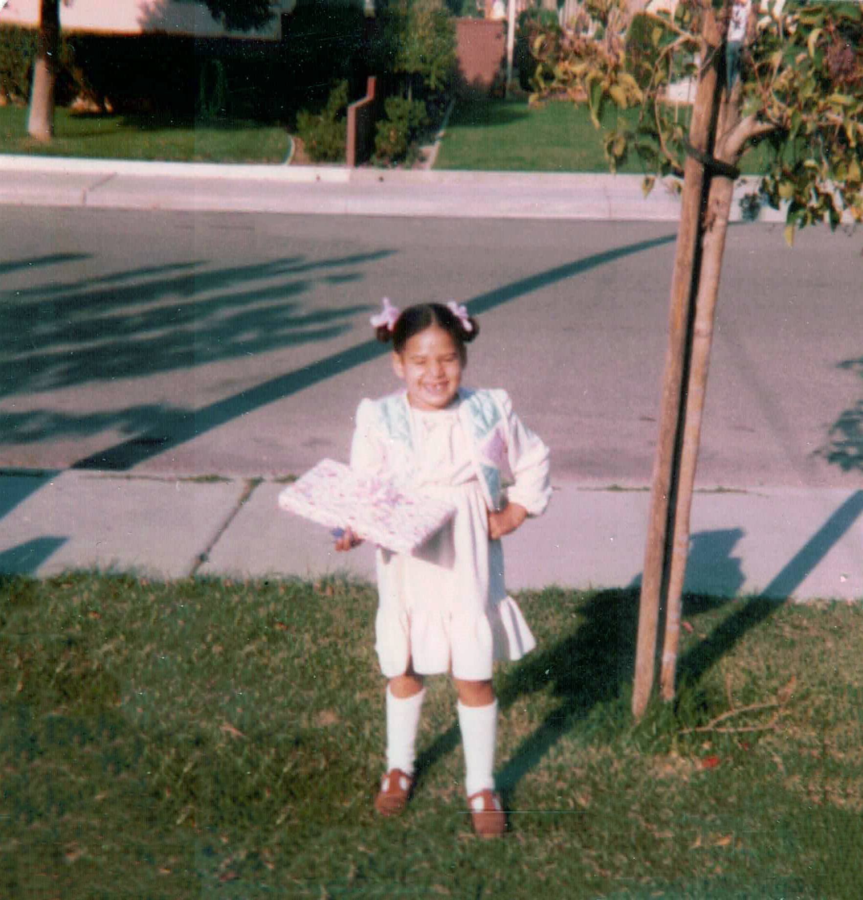 A young girl smiling while standing on a sidewalk next to a small tree in a neighborhood. She is wearing a floral dress, white knee-high socks, and brown shoes, and is holding a doll or stuffed animal. The background includes a grassy area with bushe