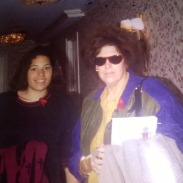 A woman and a girl standing indoors with a floral wallpaper background. The woman is wearing sunglasses and a blue and purple jacket, holding some papers, and the girl is dressed in a black top with a red bow detail.
