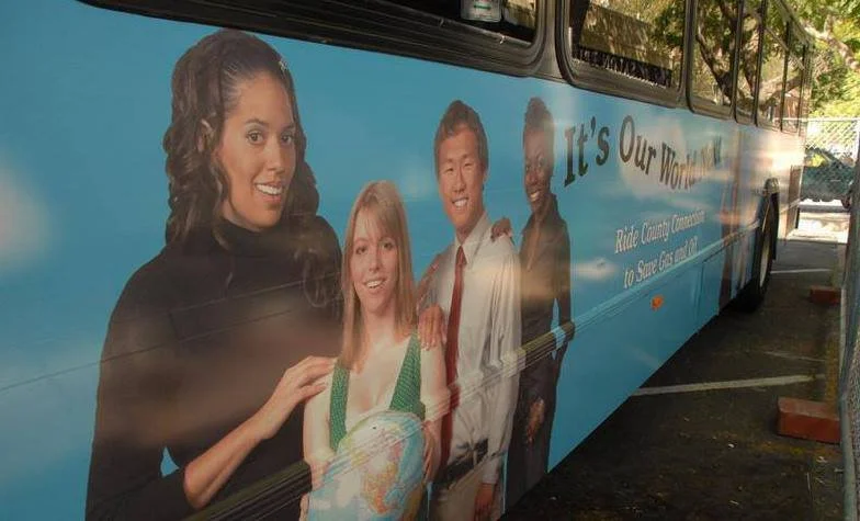 A bus with a large advertisement featuring four diverse individuals smiling, with text that reads 'It's Our World!' and 'Ride County Commuter to Serve GTS 2014'.