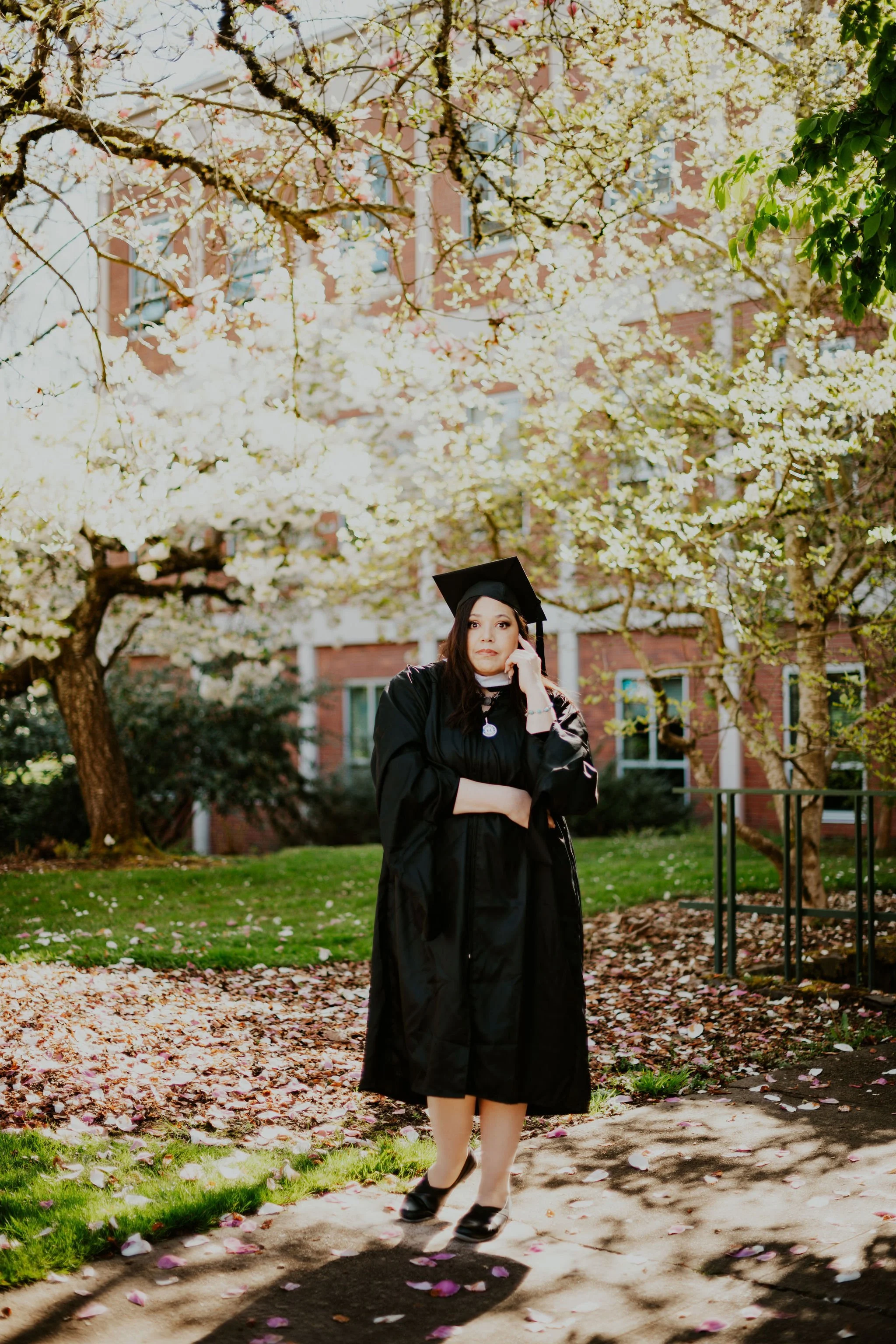 A woman in a graduation cap and gown standing outdoors in front of blossoming trees and a brick building, talking on her phone.