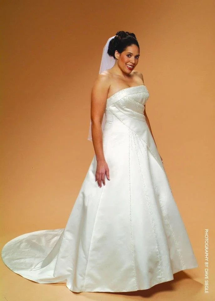 Woman in a wedding dress with a veil standing against a plain background.
