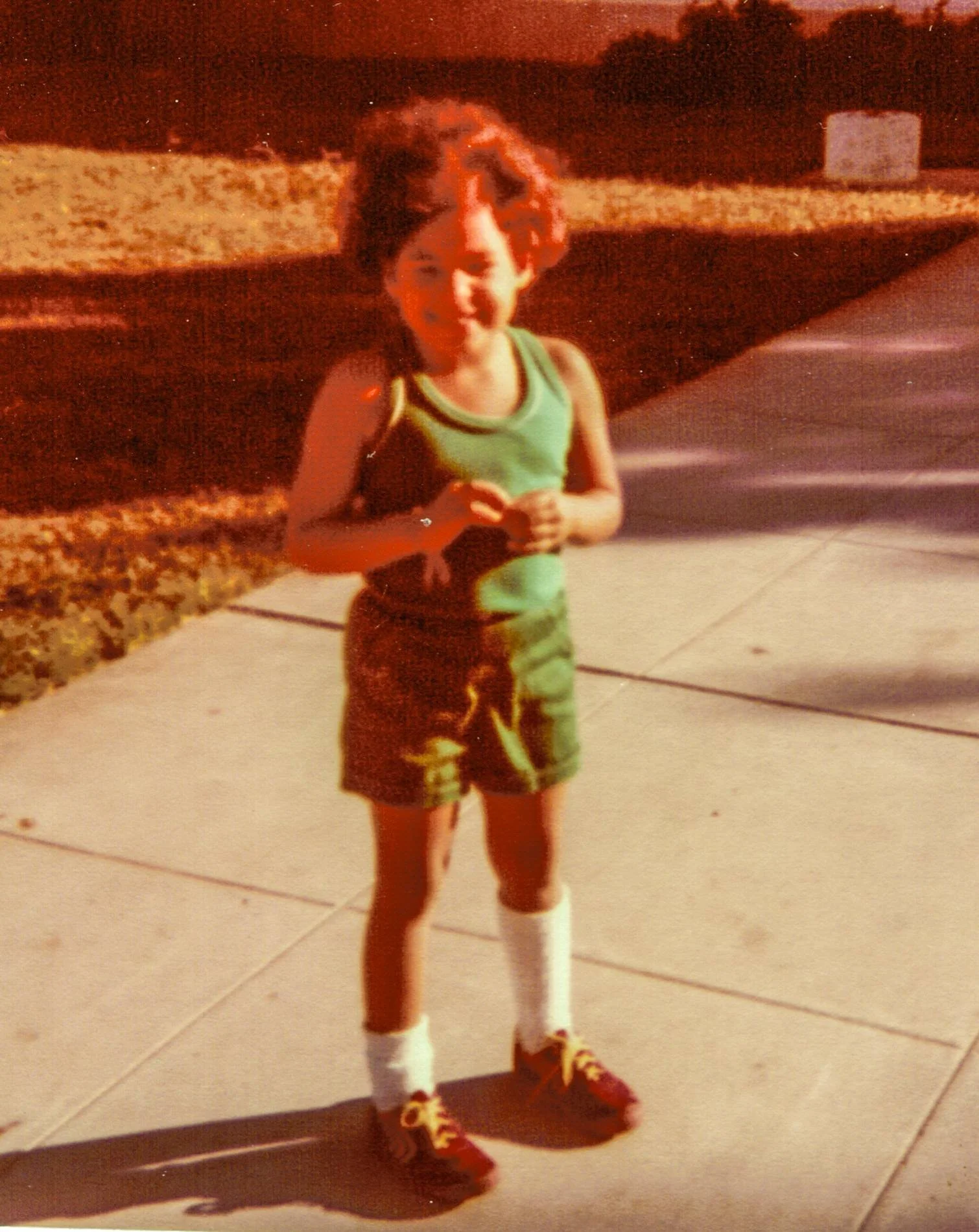A young boy standing outside at night on a sidewalk, wearing a green tank top, dark shorts, white socks, and red sneakers with yellow laces. He is smiling and has curly hair.