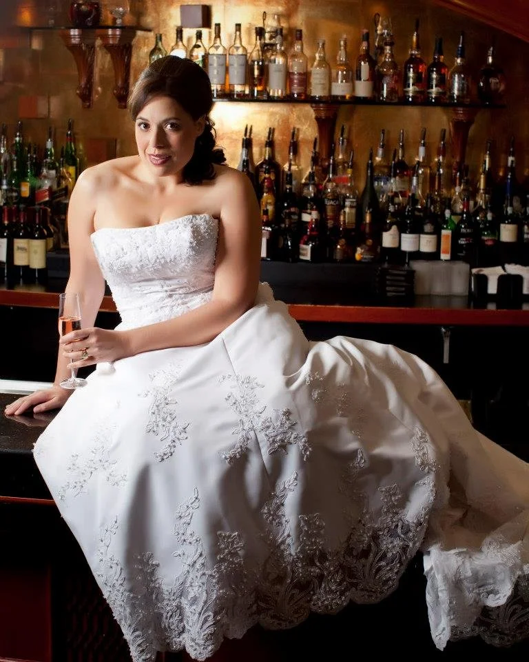 A woman in a white wedding dress sitting at a bar counter, holding a glass of champagne, with shelves of liquor bottles in the background.