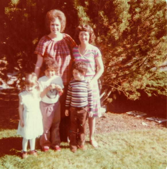 A woman and a teenage girl standing outdoors with three children, in front of a large bush or tree, on a sunny day, with a grassy area.