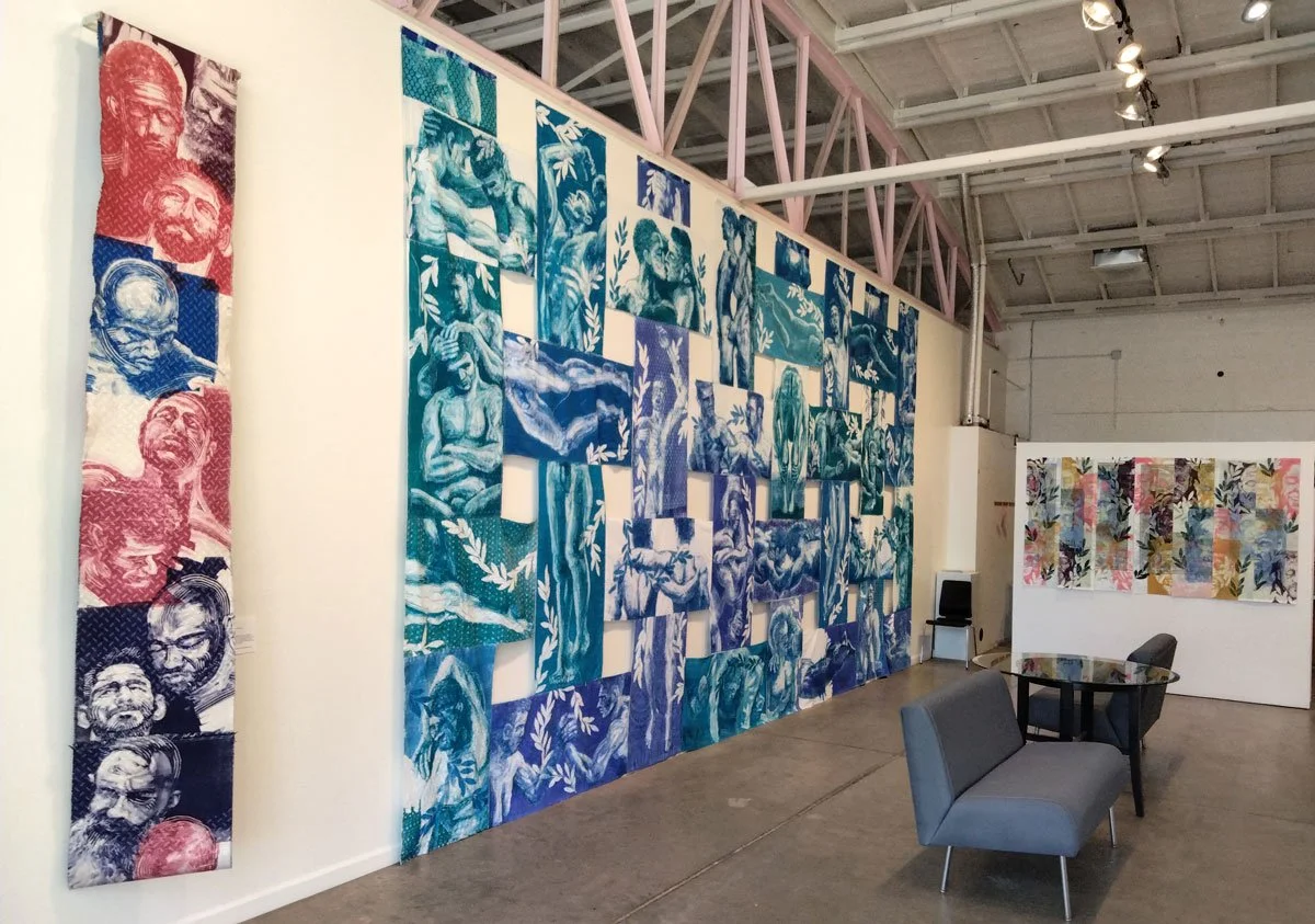 Art gallery interior featuring large textile wall hangings with various designs, including monochrome portraits and colorful abstract patterns, along with seating and tables.