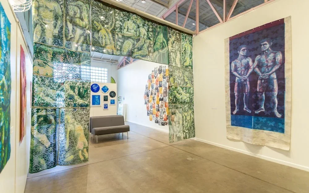 An art exhibit in a gallery featuring various textile artworks. There are fabric panels with intricate green designs forming an arch, and large fabric wall hangings with two figures on the right and a collage of smaller images in the background.