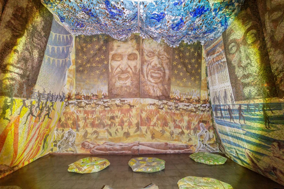 Indoor art installation with large painted murals and fabric textures. Features faces, figures, and patterns including a face with a beard on the right wall, a face with an open mouth on the back wall, and a tiger on a stand. The ceiling and walls are decorated with abstract, colorful patterns.