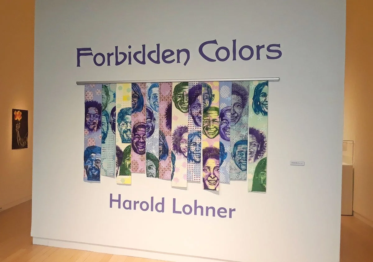 Artwork titled "Forbidden Colors" by Harold Lohner, featuring multiple portraits of smiling faces in vibrant colors displayed on hanging panels.
