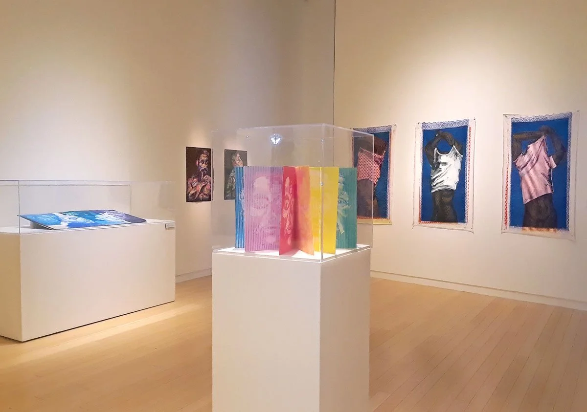 An art gallery with various framed photographs of people on the wall, a colorful book display in a glass case at the center, and a white display table with magazines on it.