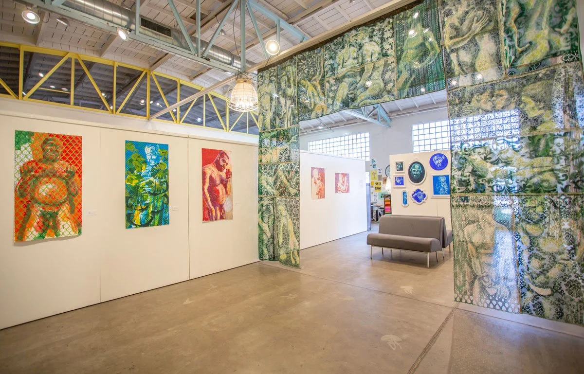 Art gallery with colorful paintings of human figures on white walls and decorative fabric panels with a nature motif hanging overhead. A gray sofa is placed near the artwork, and the space has high ceilings with metal beams and large windows.