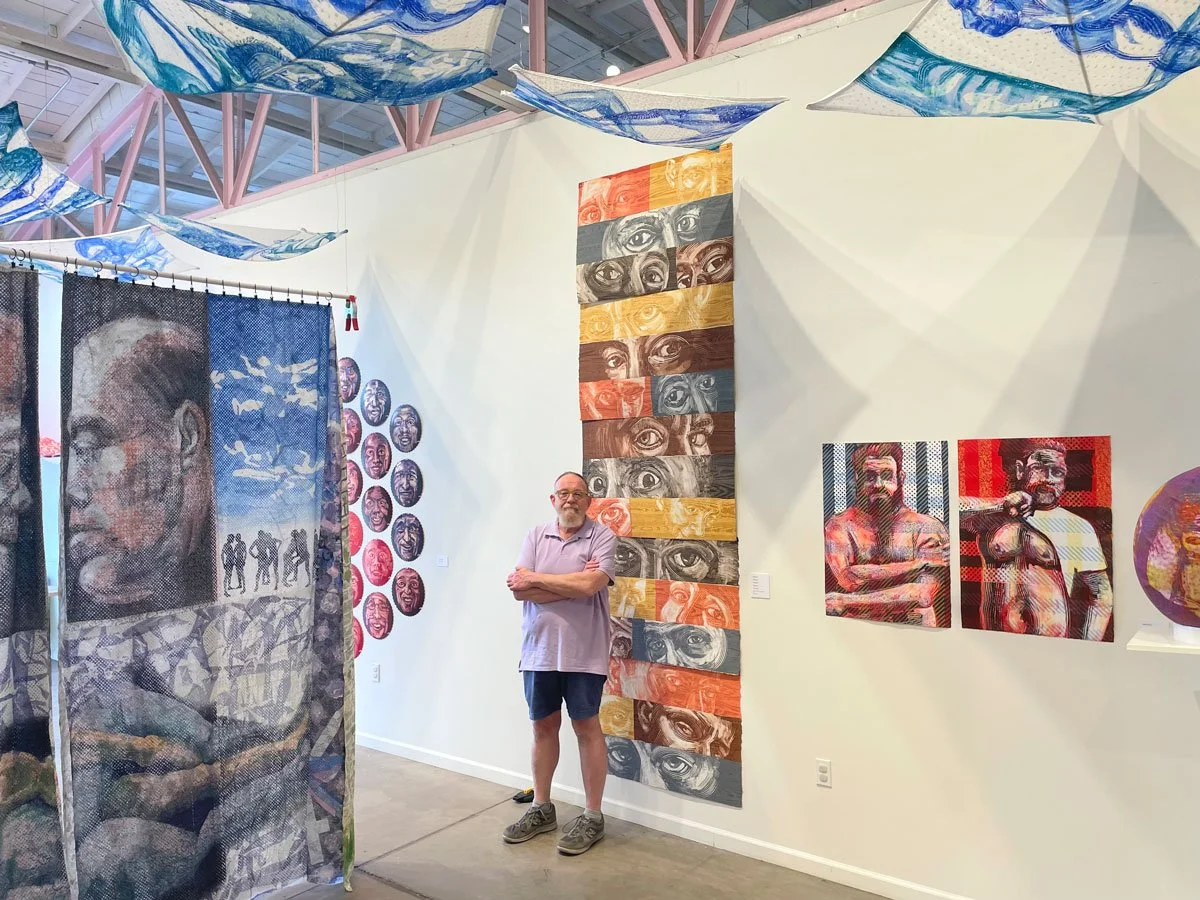 An art gallery with colorful portraits and abstract pieces. A man stands with arms crossed next to a vertical panel of painted eyes, with other paintings of faces and eye motifs around him.