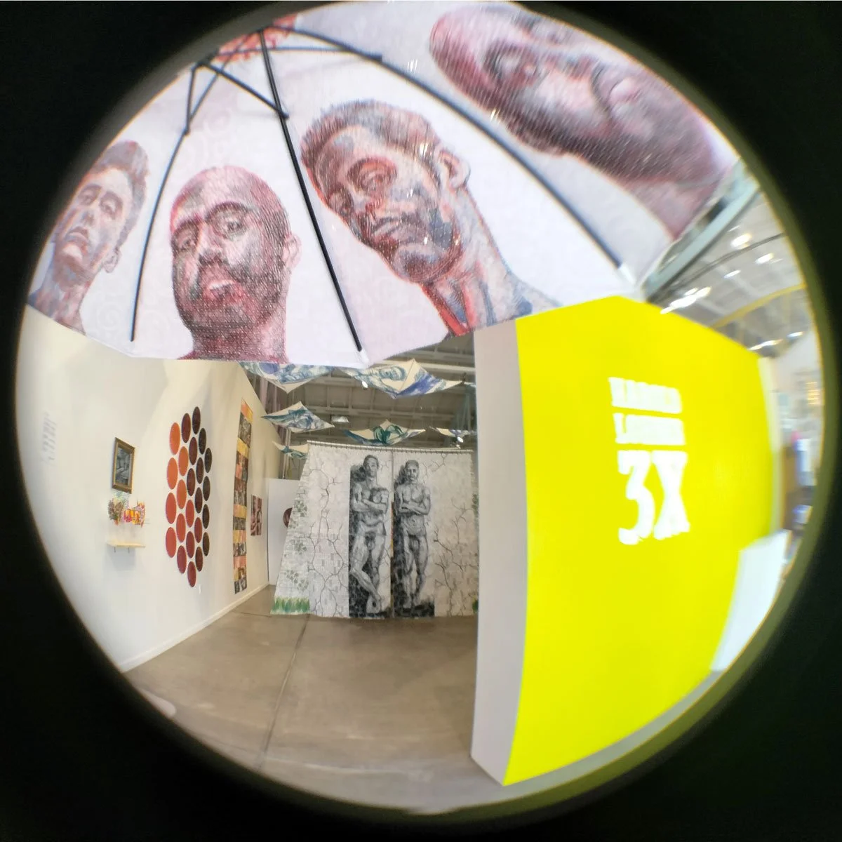An art exhibit featuring portraits and sculptures, including a large umbrella with urban street art-style faces, a drawing of two men with crosses on the wall, and yellow and white signage.