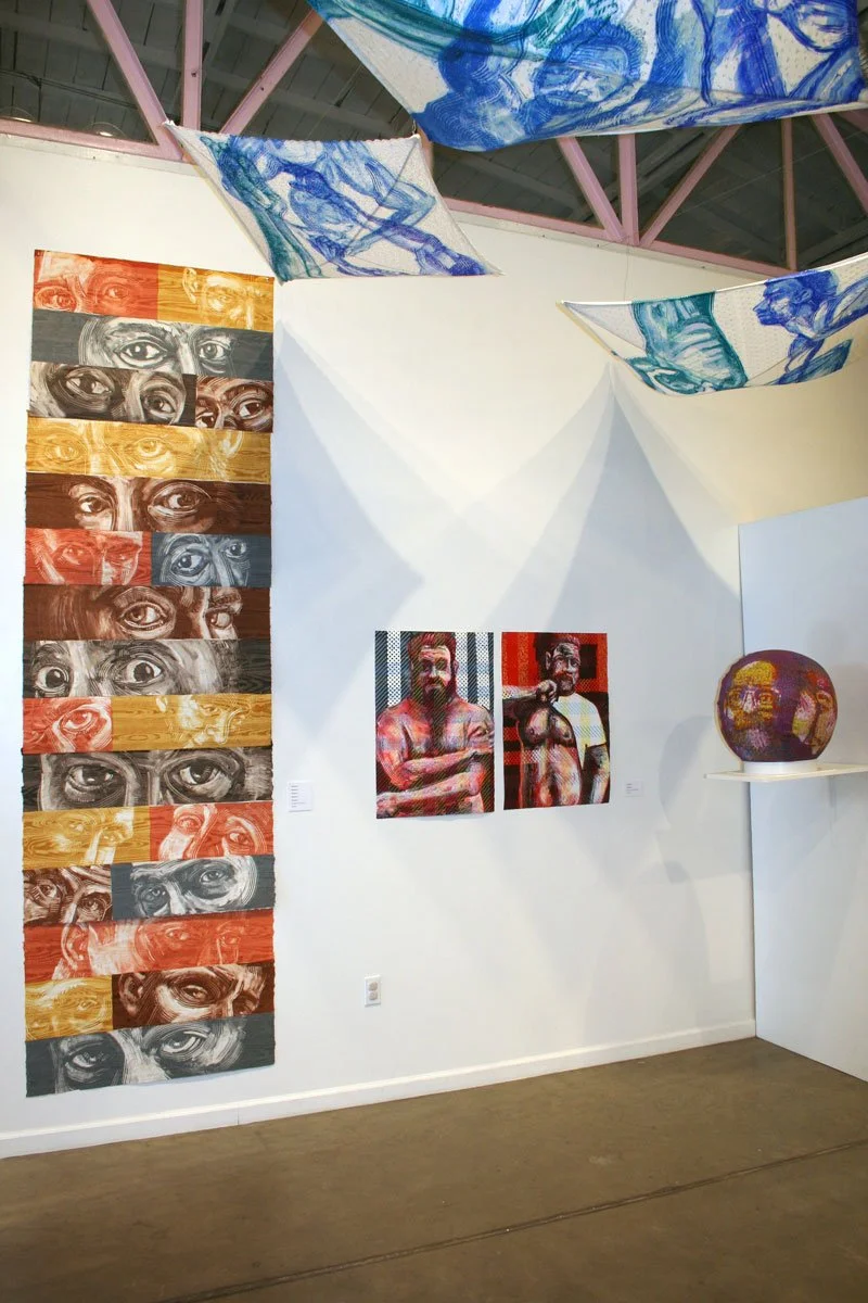 Art exhibition featuring a vertical multi-panel piece of eyes in black, white, red, orange, gray, and yellow tones; two paintings of shirtless men with beards and tattoos; a spherical sculpture on a white shelf; and fabric art pieces hanging from the