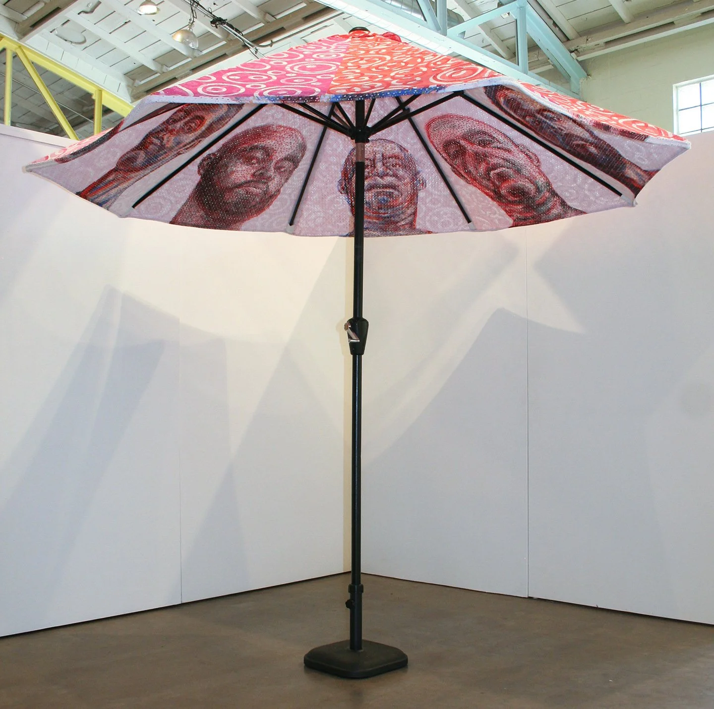 A large umbrella with portraits of three people's faces painted on the inside, displayed indoors against a white wall.