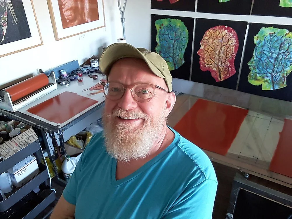 A man with a beard and glasses taking a selfie in an art studio, with colorful abstract paintings of human profiles and brain illustrations on the walls, and art supplies on tables.