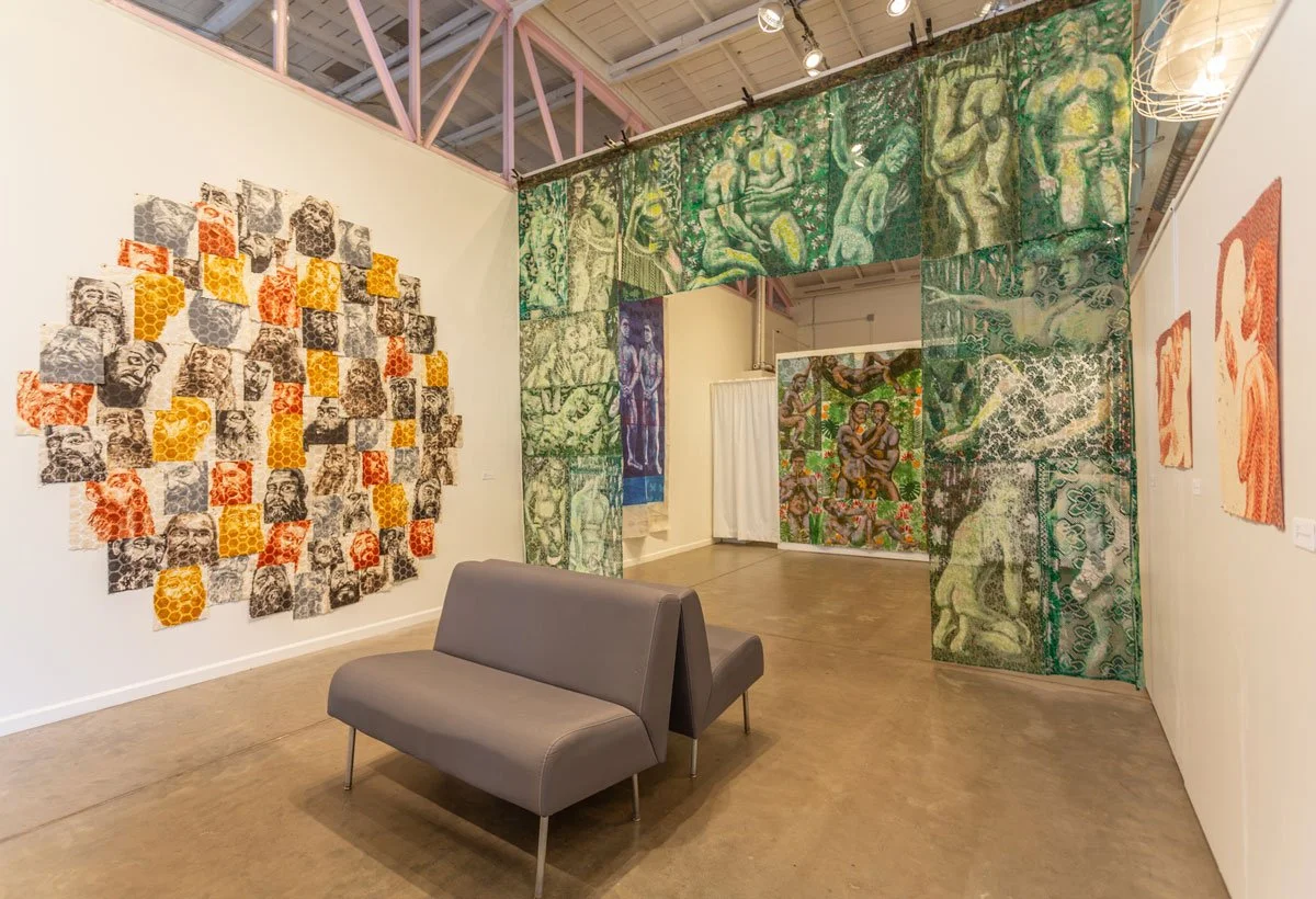 Art gallery with various colorful abstract and figurative paintings on the walls and hanging fabric. A gray bench is in the foreground, and track lighting illuminates the artwork.