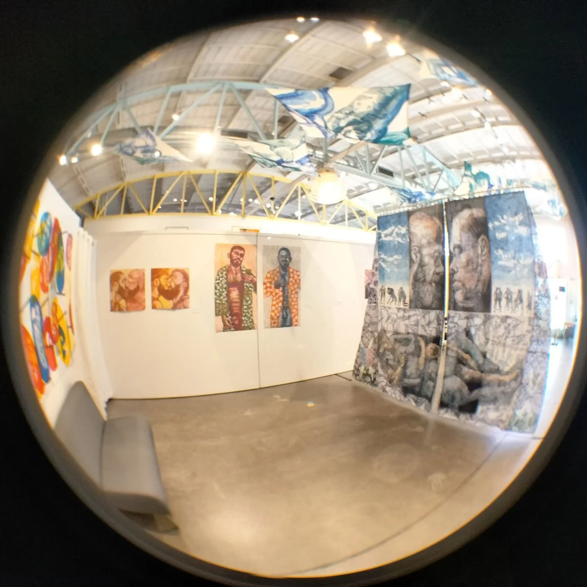 Art gallery with various paintings and murals on display, featuring portraits, abstract designs, and mixed media artworks, with a curved, metallic ceiling and hanging fabric art pieces.
