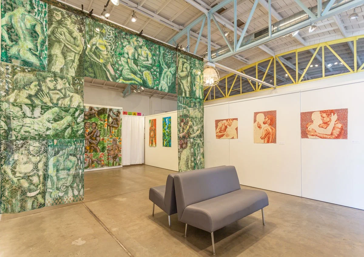 An art gallery with murals and paintings, including green-toned murals on a curved wall and pastel-colored portraits on white walls, with a gray sofa in the foreground and a high ceiling with metal beams.
