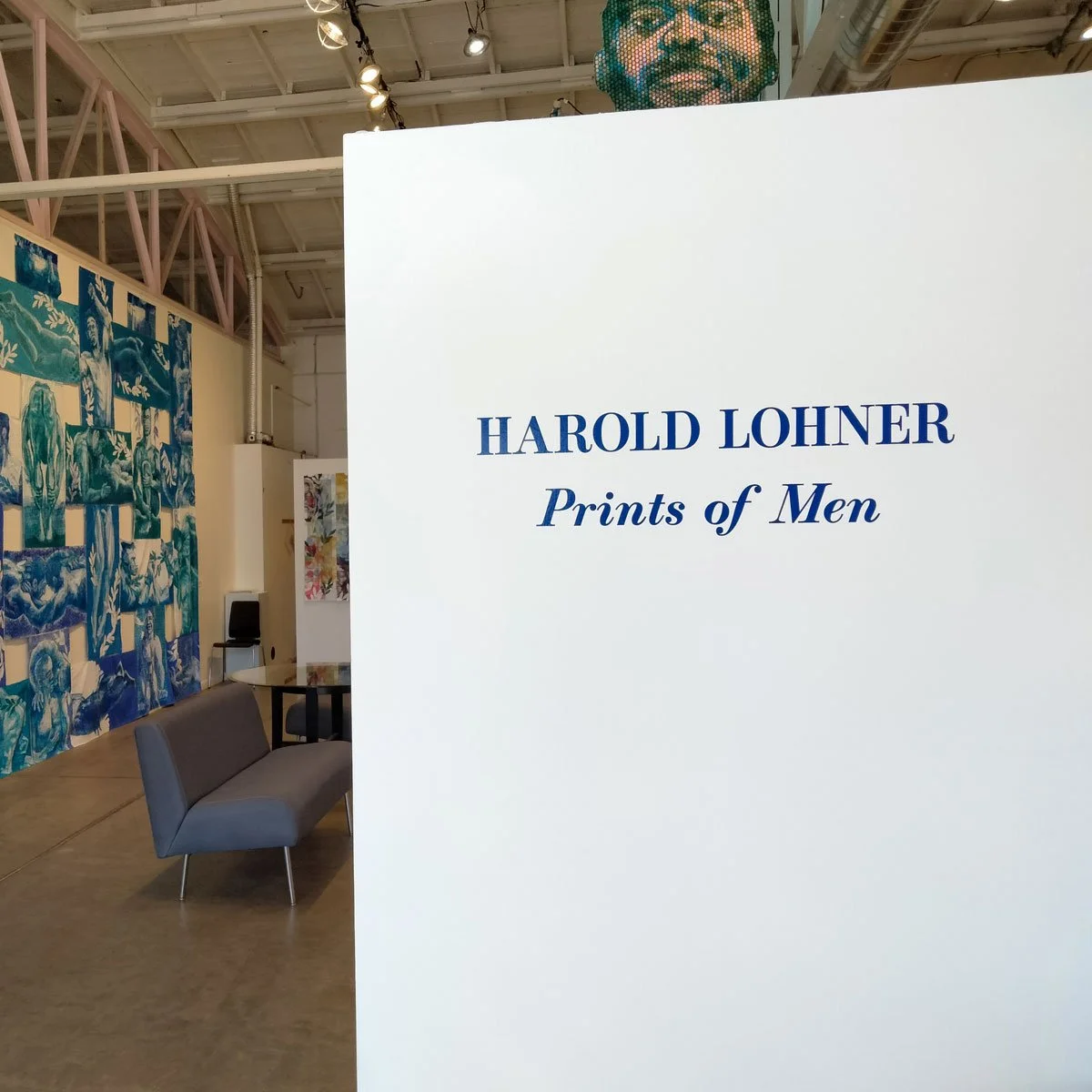 Gallery sign reading 'Harold Lohner Prints of Men' with artwork and seating area visible in the background.