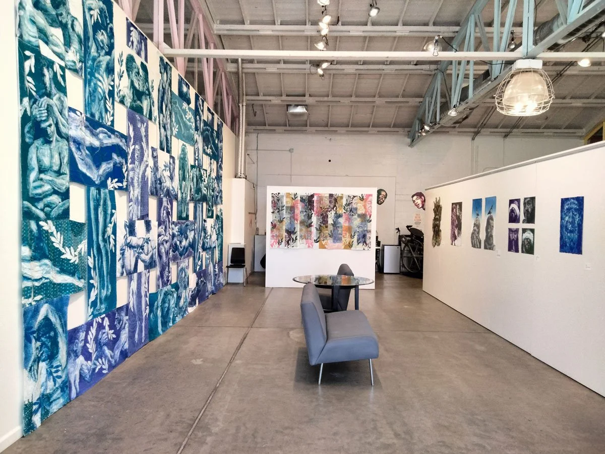Art gallery featuring various paintings and mixed media artwork displayed on white walls inside a spacious industrial-style building.