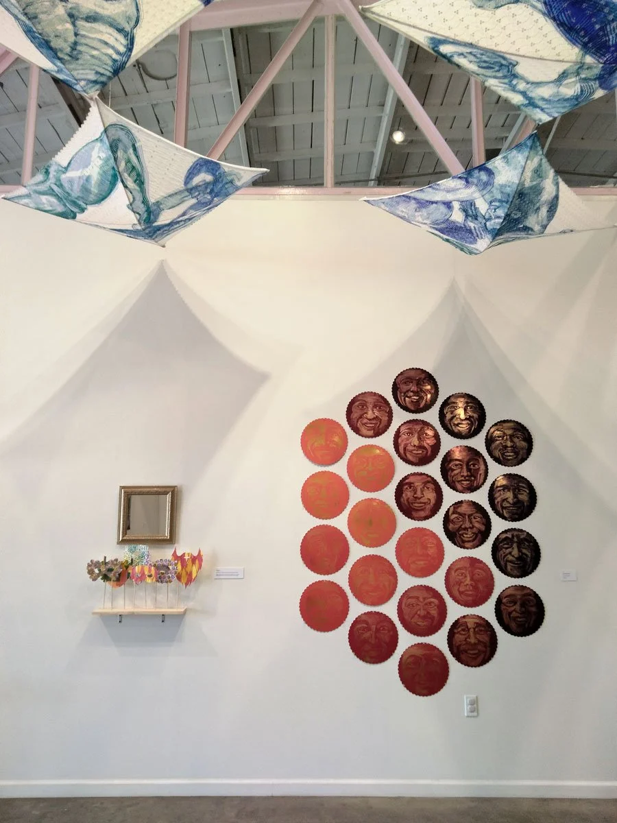 Contemporary art installation with multiple circular portraits of a smiling person's face arranged in a pattern on a white wall, and a small shelf holding colorful paper-crafted flowers, with a decorative empty frame above it, and blue and white fabr