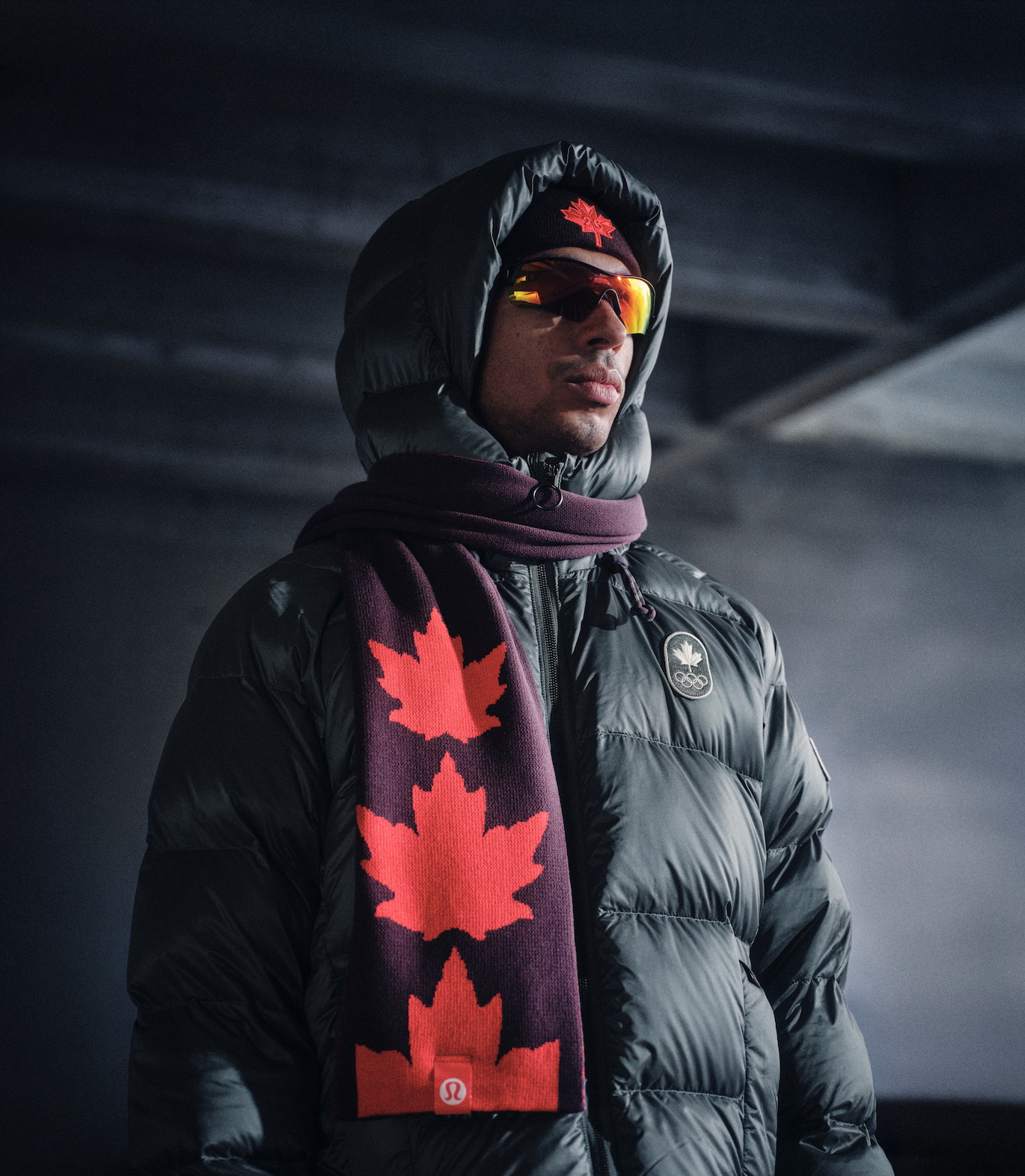 A male athlete wearing a black puffer jacket, a black beanie with a red maple leaf logo, reflective sunglasses, and a maroon scarf with red maple leaves, standing indoors in a dimly lit space.