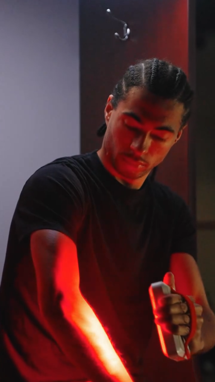 A man with braided hair looking at his arm illuminated in red light while holding a white object, possibly a phone, in his right hand.