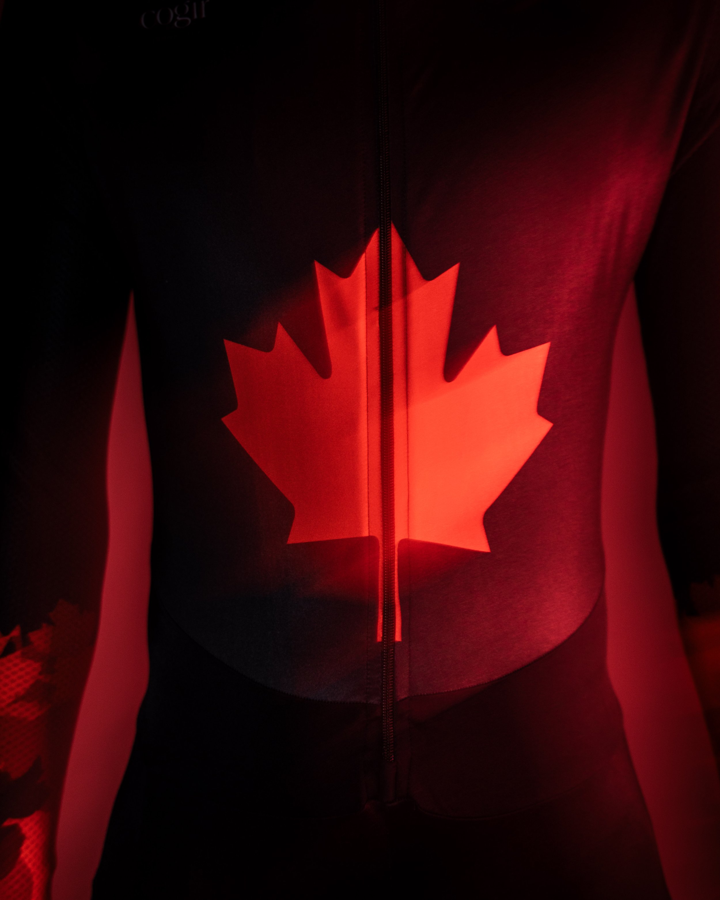 A person wearing a black zip-up jacket with a bright red Canadian flag design on the front, illuminated by red light.