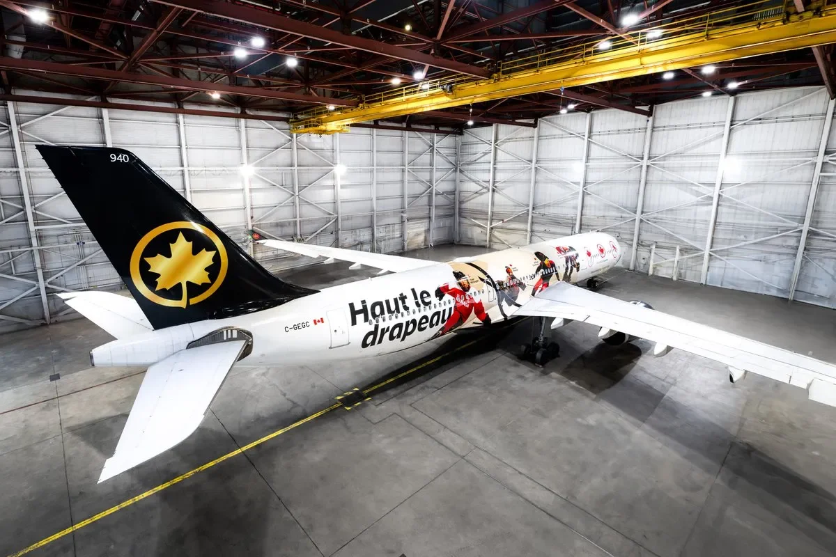 Inside a hangar, a large airplane with a Canadian flag is being painted with a festive holiday theme featuring Santa Claus. The plane's tail features a black and gold maple leaf logo. The aircraft has text that reads 'Haute le drapeau' on its fuselage.
