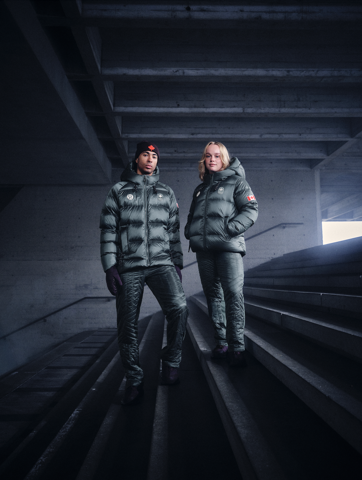 Two athletes in dark green, insulated jackets standing on stairs in a concrete structure, with one woman on the right and one man on the left, both looking at the camera.