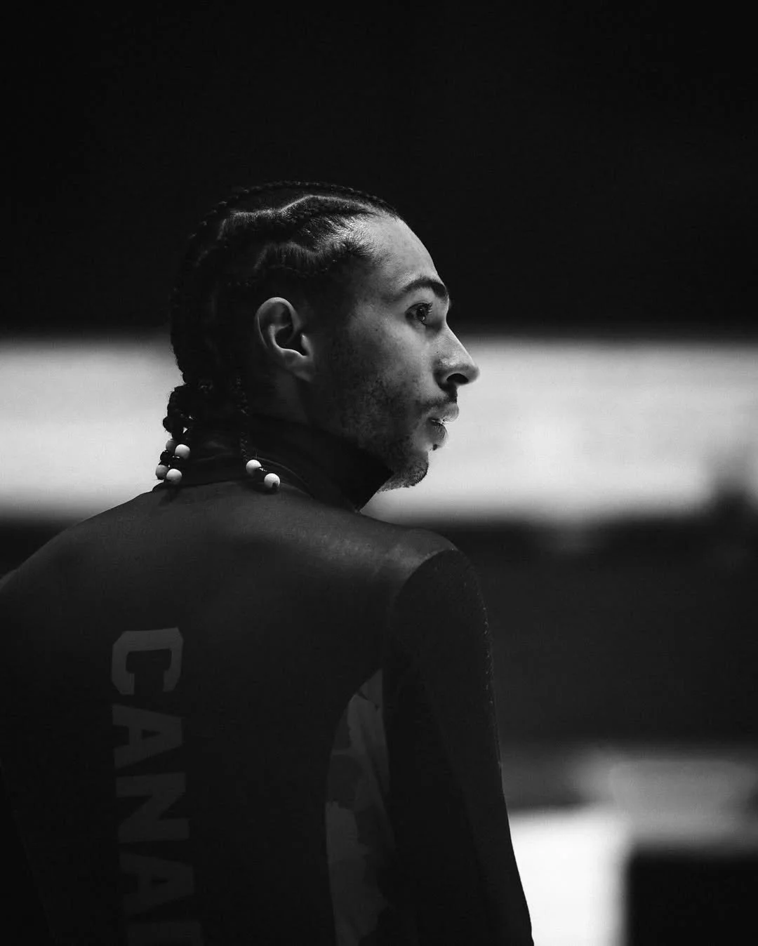 Black and white side profile of an athlete with braided hair, wearing a jersey with 'CANADA' on the back, sitting and looking to the right.