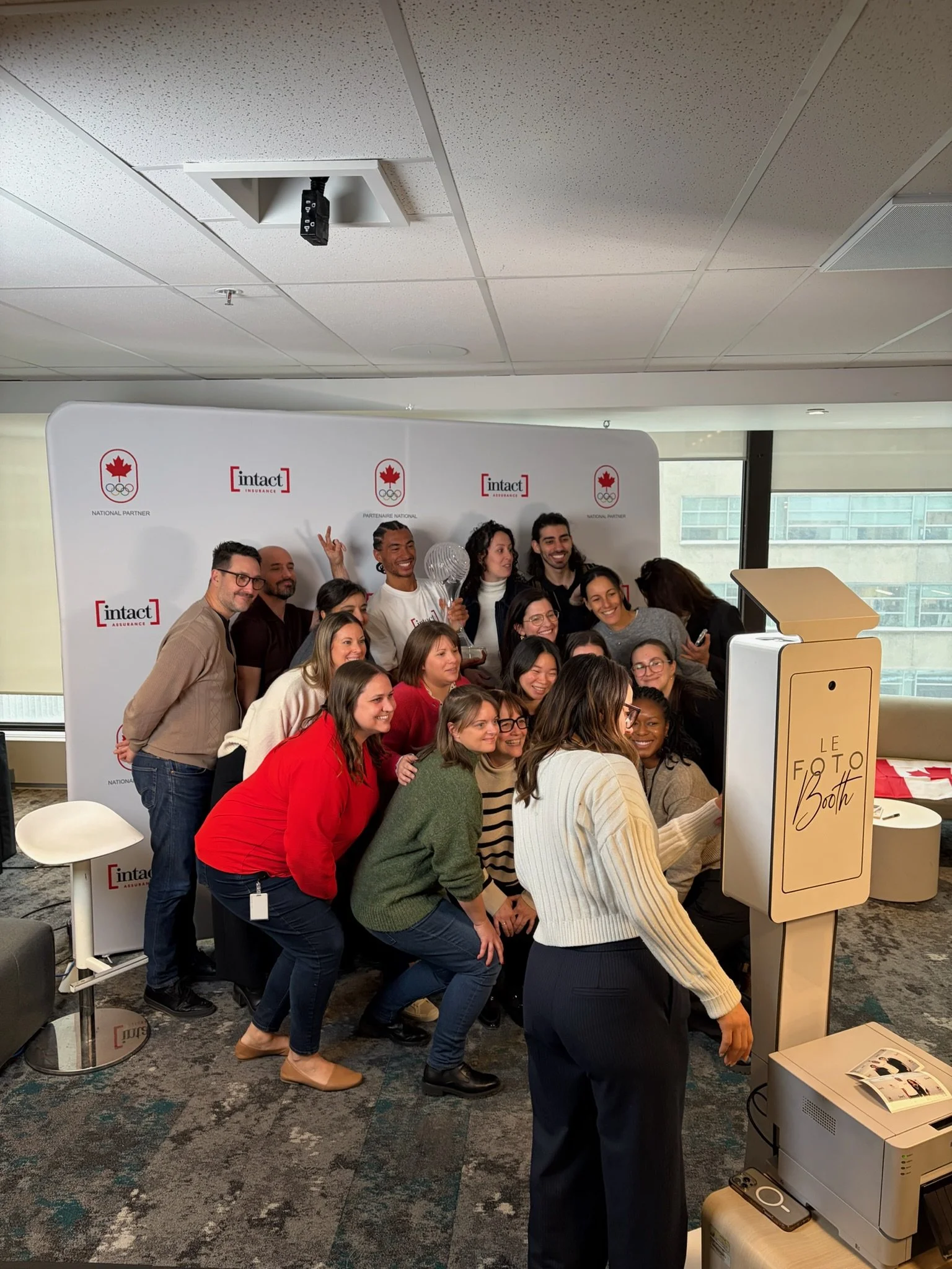A group of people gathered in front of a photo booth at a corporate event, smiling and posing for a picture. The background has a white banner with logos and text.