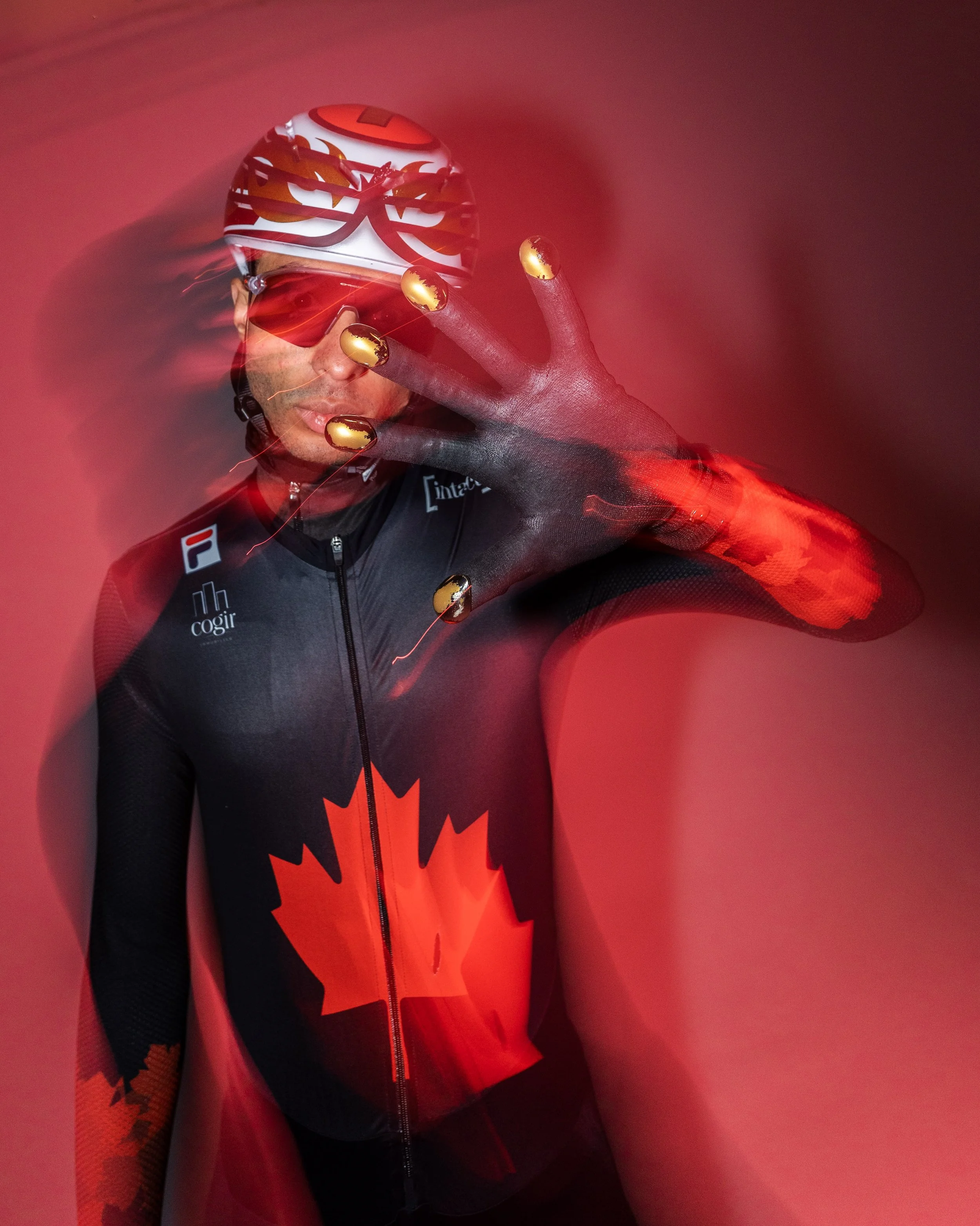 A cyclist wearing a maple leaf design cycling suit and helmet, holding his gloved hand in front of the camera with a blurred red background.