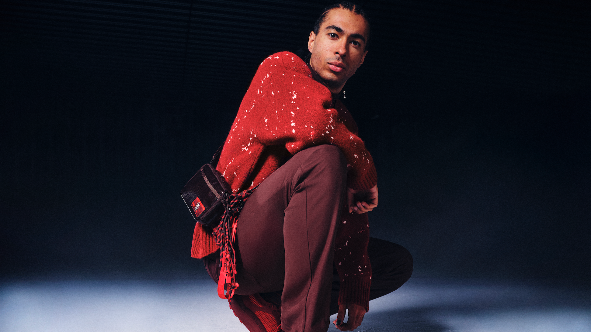 A man with dark hair and a light skin tone wearing a red sweater with white specks and brown pants, sitting in a dark studio setting with a gradient background.