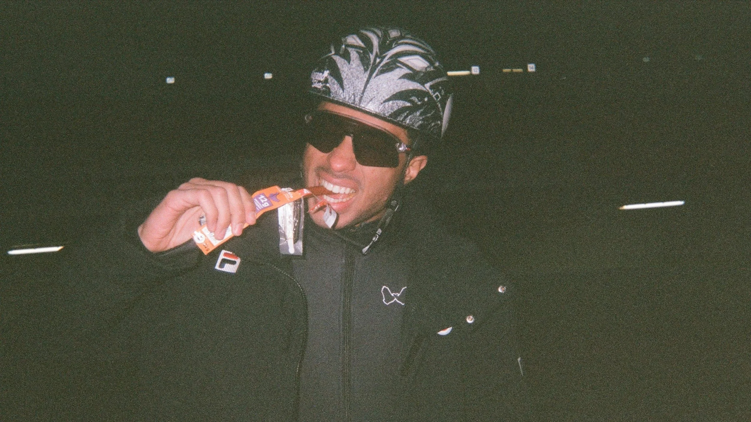 A person in cycling gear, including a patterned helmet and sunglasses, is biting into a snack bar while holding it in their hand. The person is smiling and appears to be in a dark environment, possibly after a cycling session.