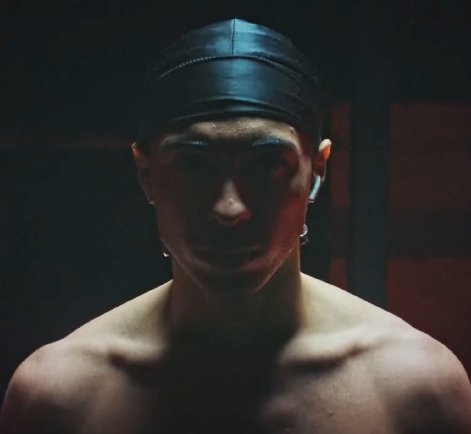 A young man with a black durag and AirPods, shirtless, standing in a dark room with red lighting, facing forward.