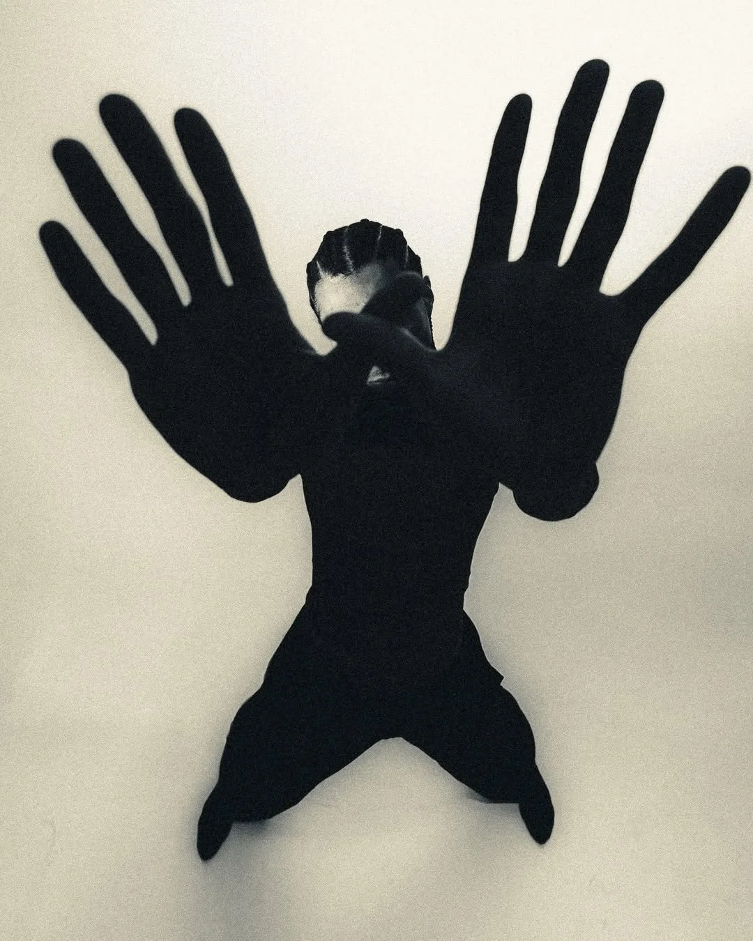 A black cutout of a person's hands and body on a white wall, with the actual person standing behind it, facing the camera, creating a layered visual effect.