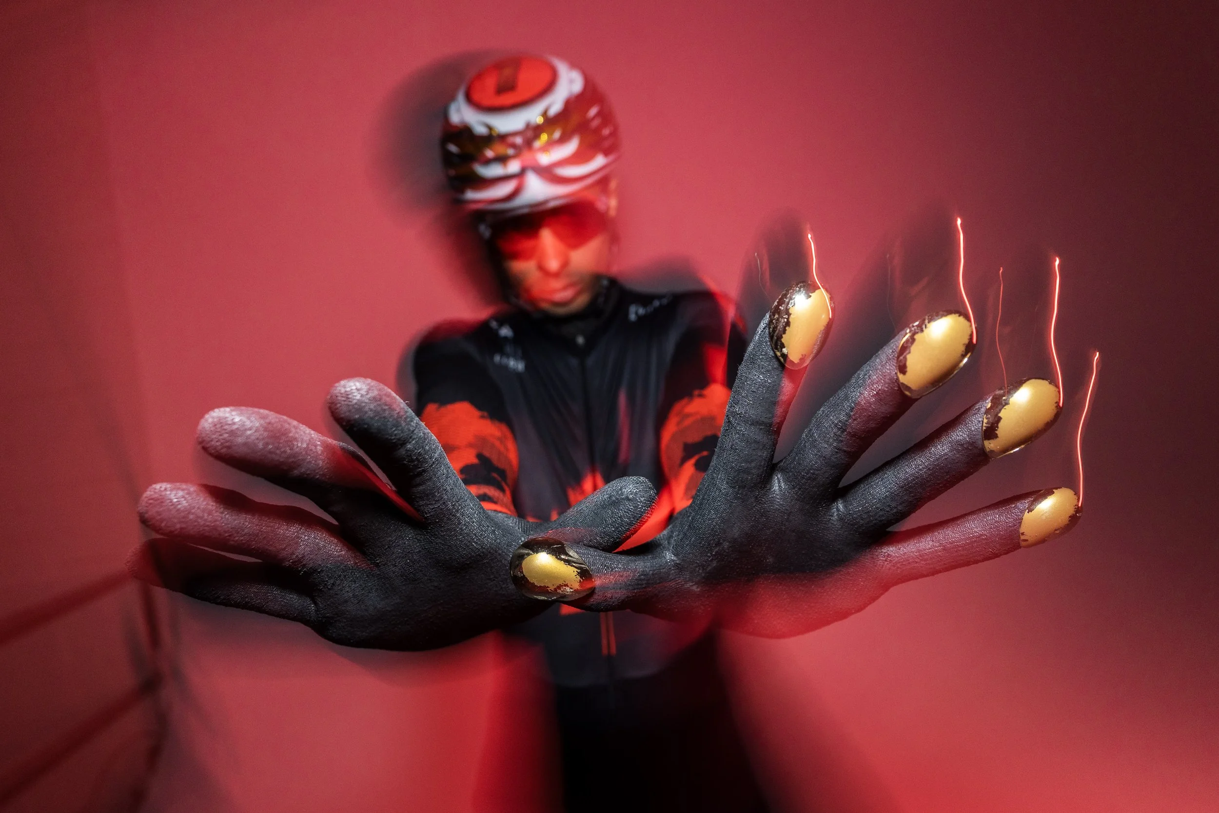 A person wearing a bike helmet, sunglasses, and sportswear, reaching out with a gloved hand, with sparks flying from their fingers, against a red background.