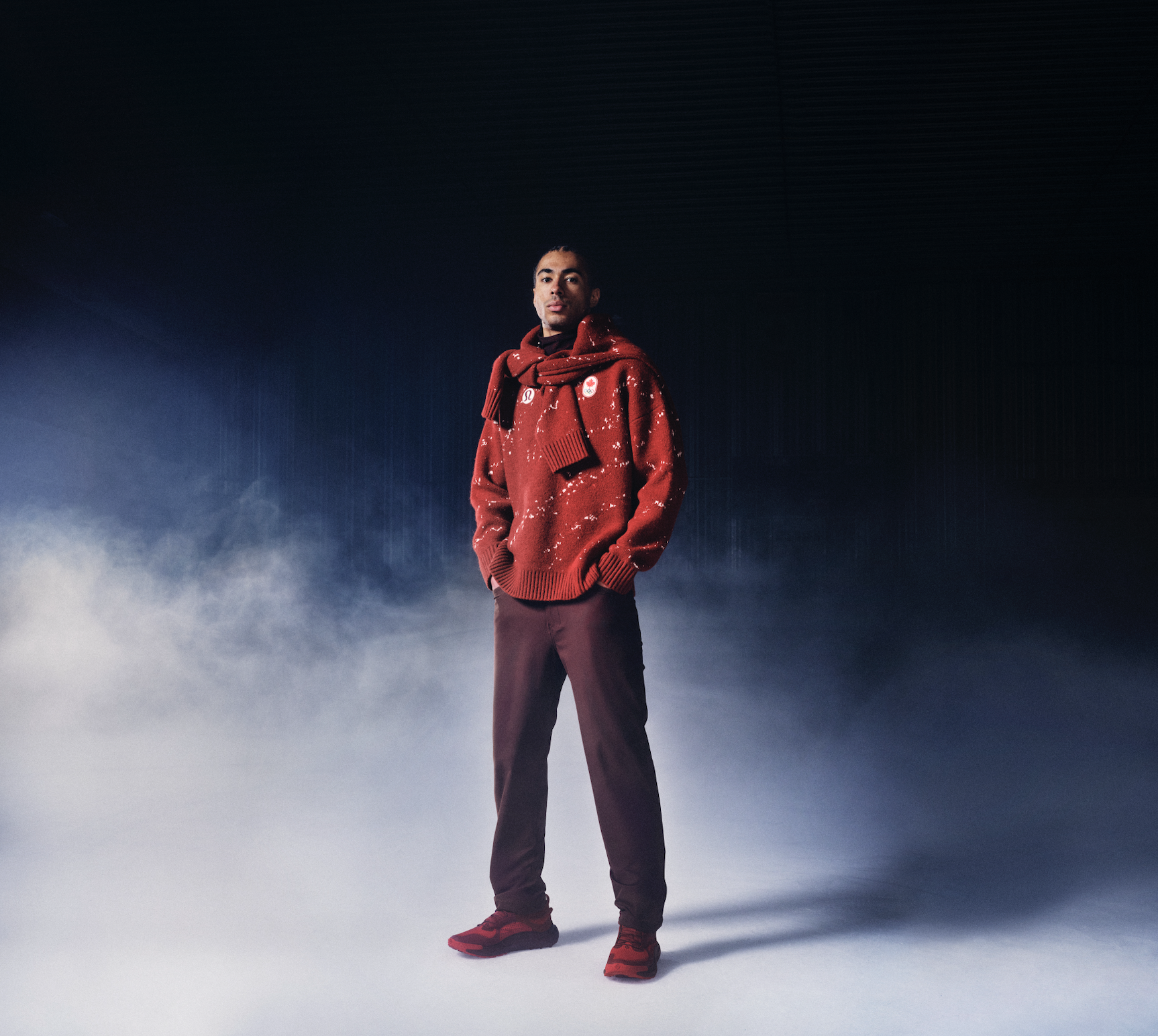 A man in a red sweater with snowflake patterns, red sneakers, and dark pants stands in a misty or foggy environment with a dark, textured background.