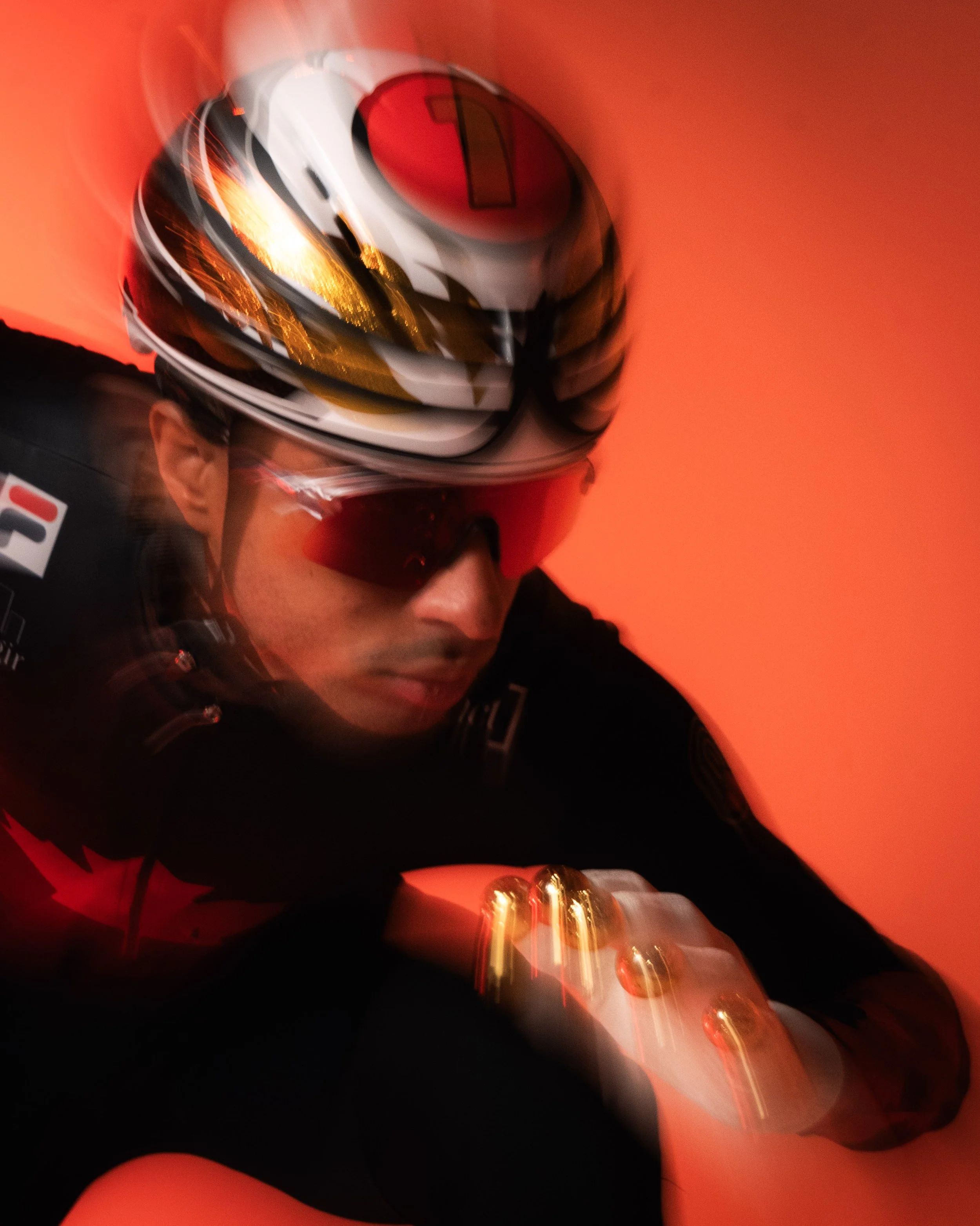A person wearing a racing helmet with a black, white, and gold design, and a racing suit, looking down with a red background, possibly in a racing environment.