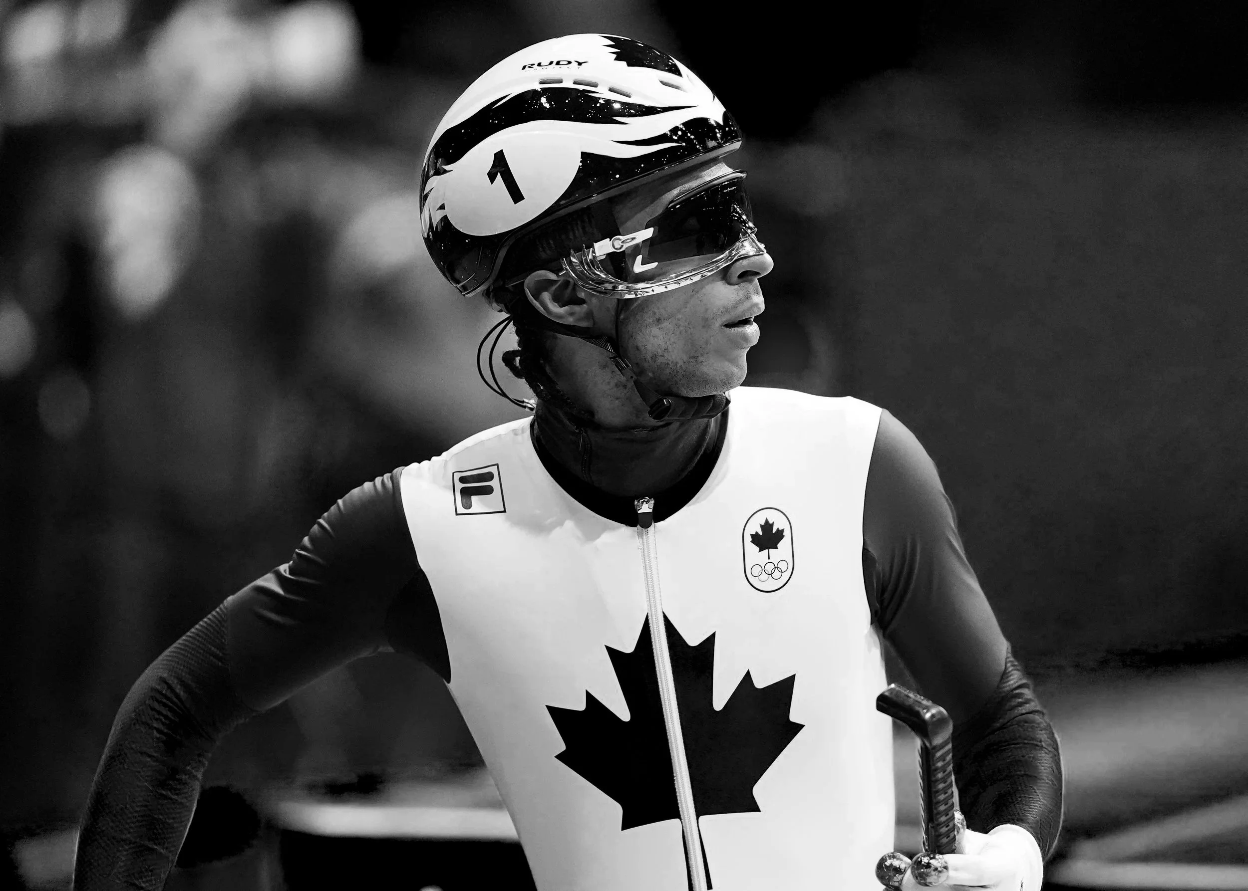 A female athlete in a athletic uniform with a Canadian emblem and the Olympic rings, wearing a helmet and goggles, standing with her hands on her hips in a black and white photo.