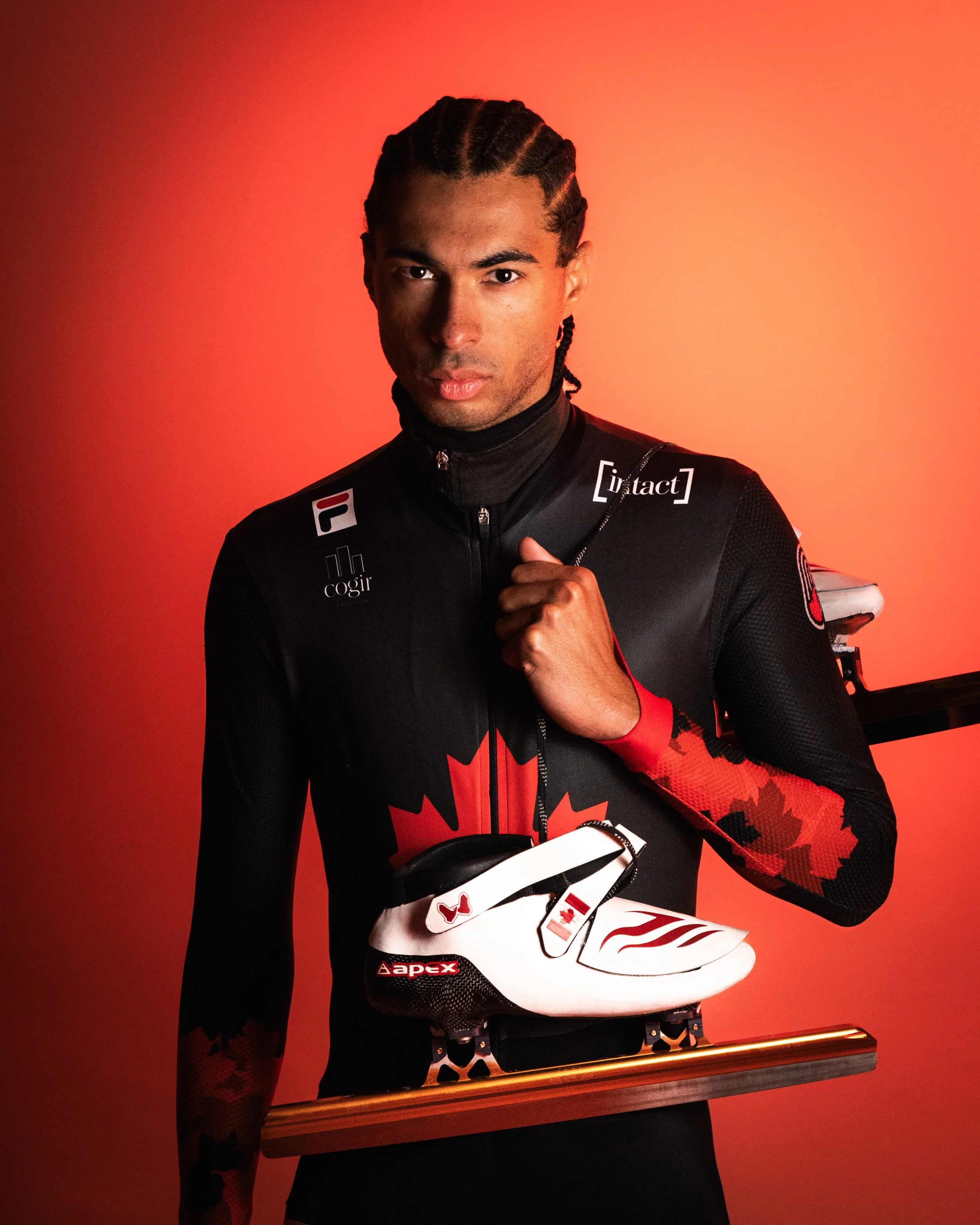 A person dressed in black and red cycling gear holding a white cycling shoe and a wooden stick against a red background.