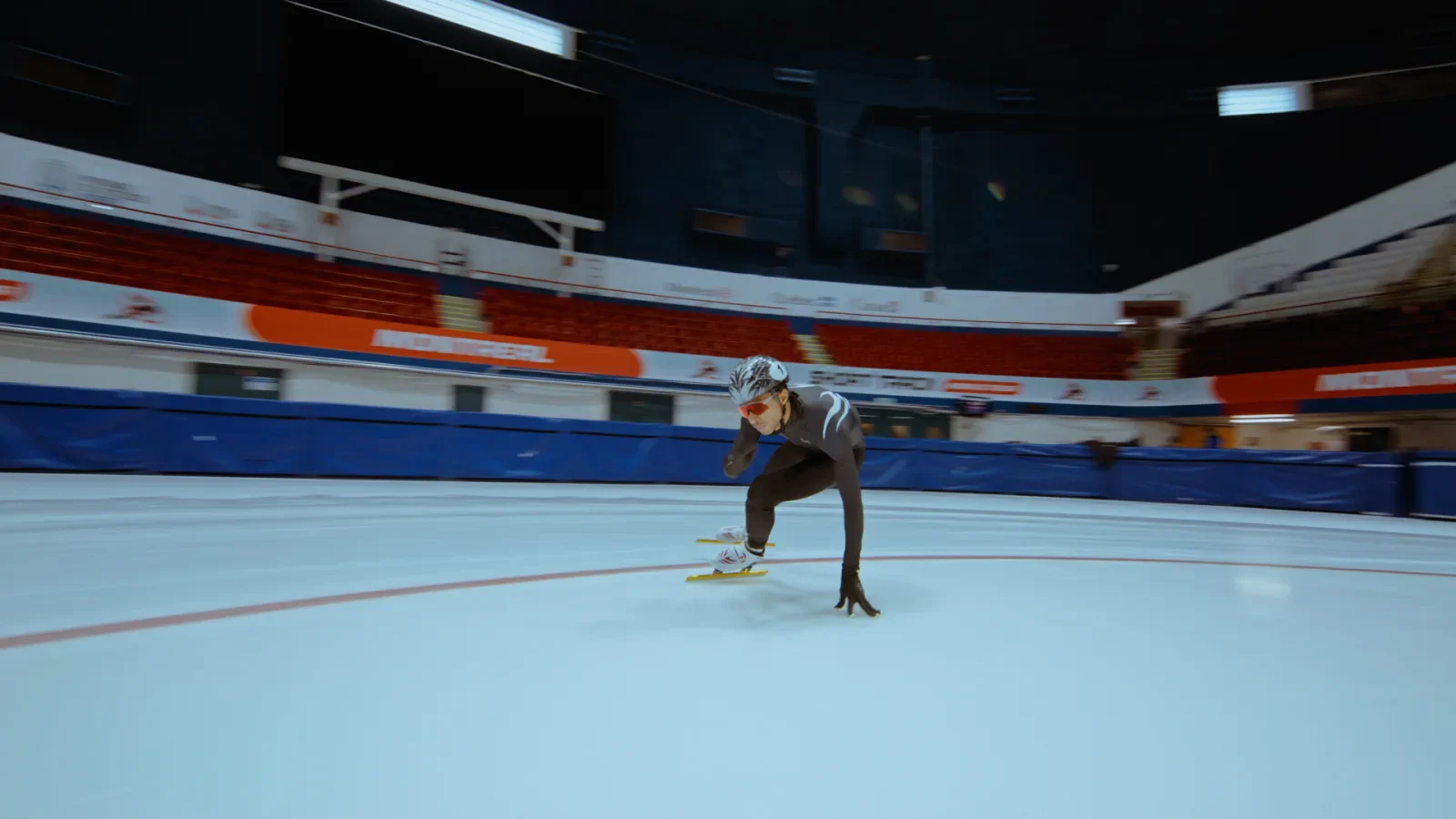 A person in an indoor ice skating rink, dressed in a black bodysuit, helmet, goggles, and gloves, is skating rapidly on the ice, leaning forward with one hand touching the ice.