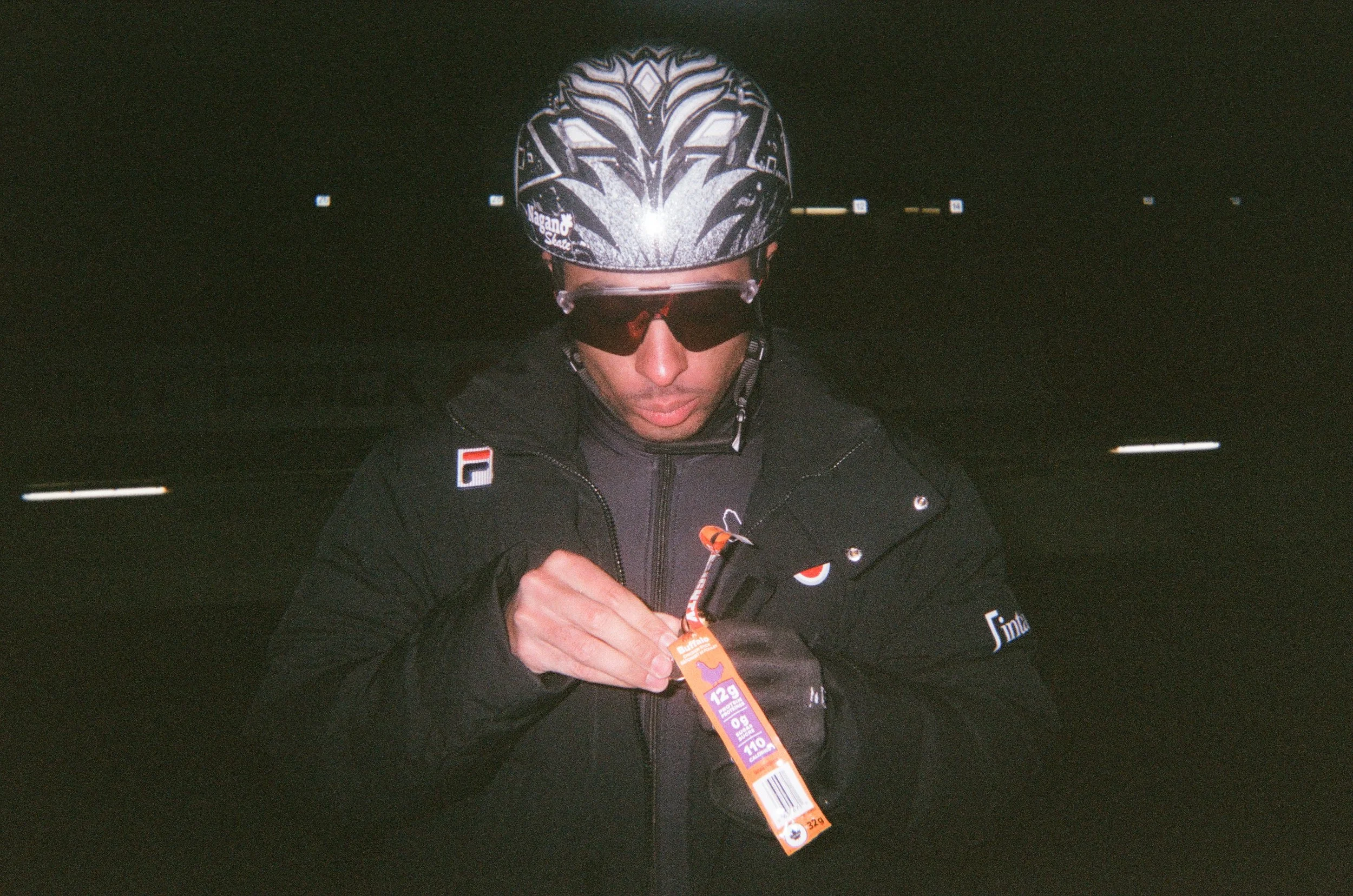 A person wearing a black cycling helmet with white and black designs, black sunglasses, and a black jacket with various logos, looking down at a runner's bib with race information.