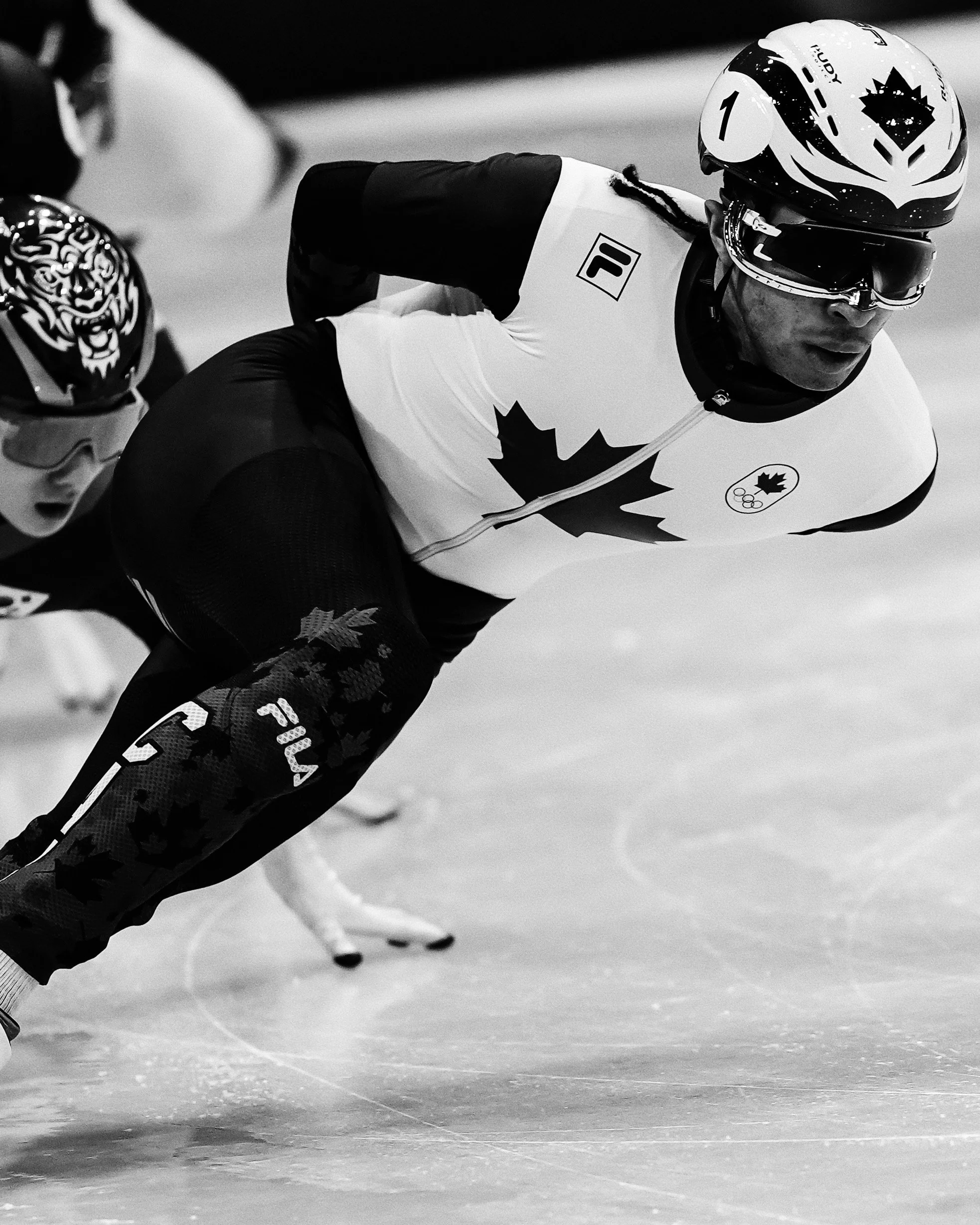 A speed skater leans into a turn on the ice, wearing a helmet, goggles, a fitted racing suit with Canada and Olympic logos, and skates with curved blades.