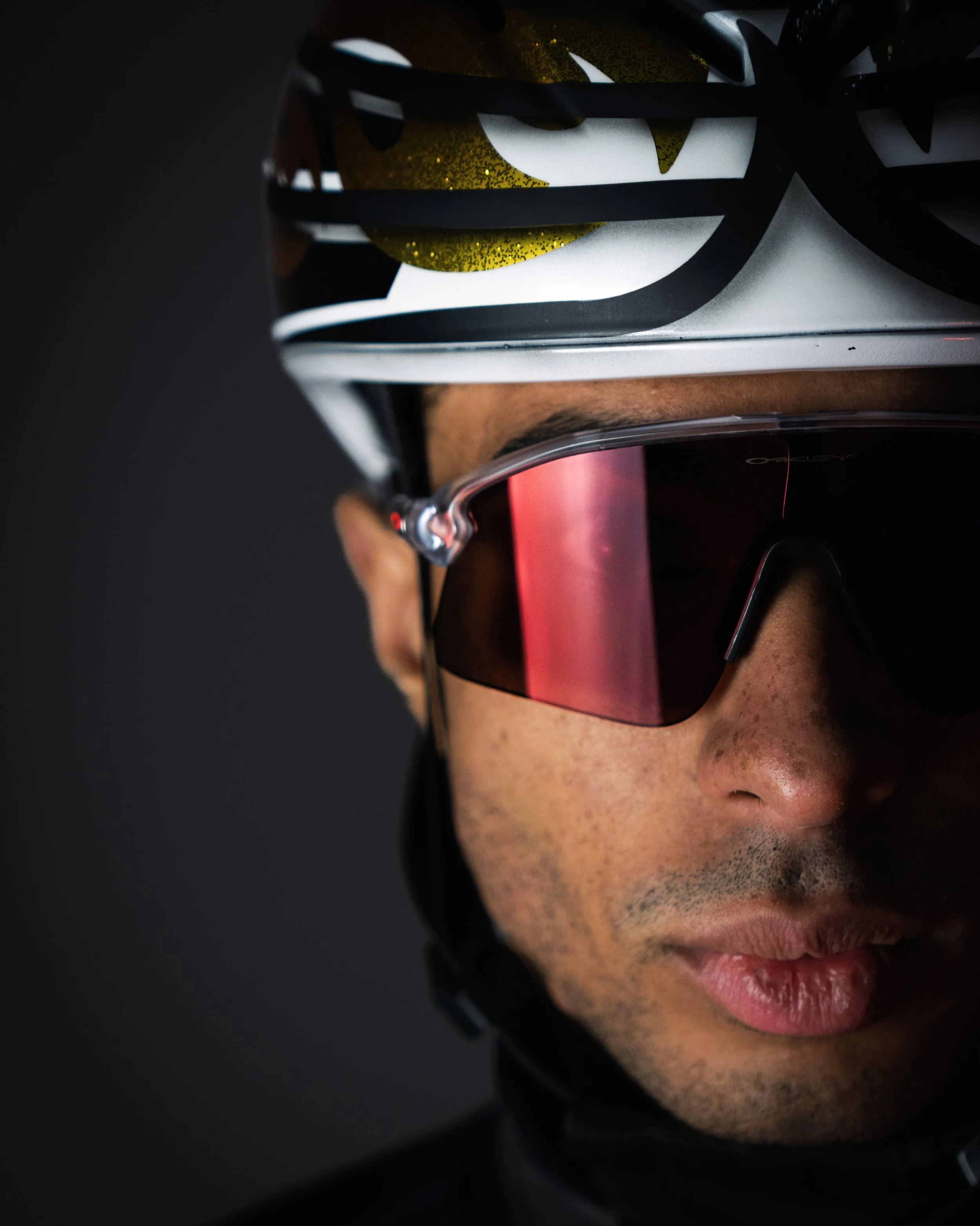 Close-up of a man wearing a Nike cap, sunglasses with a pink reflective lens, black headphones, and red lipstick, with a dark background.